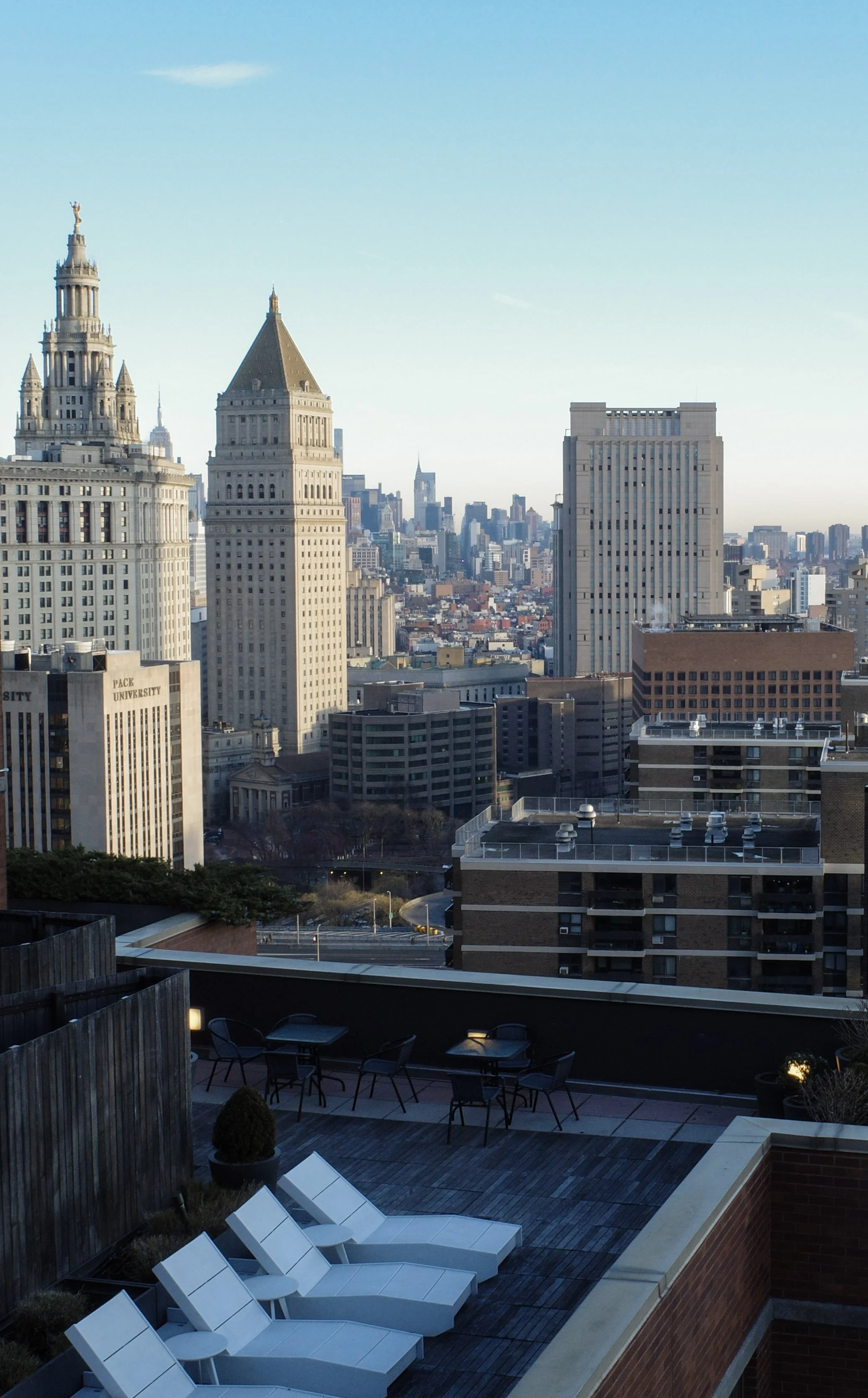 A city skyline featuring tall buildings with the Empire State Building visible in the distance, seen from a rooftop terrace with chairs and lounge chairs.
