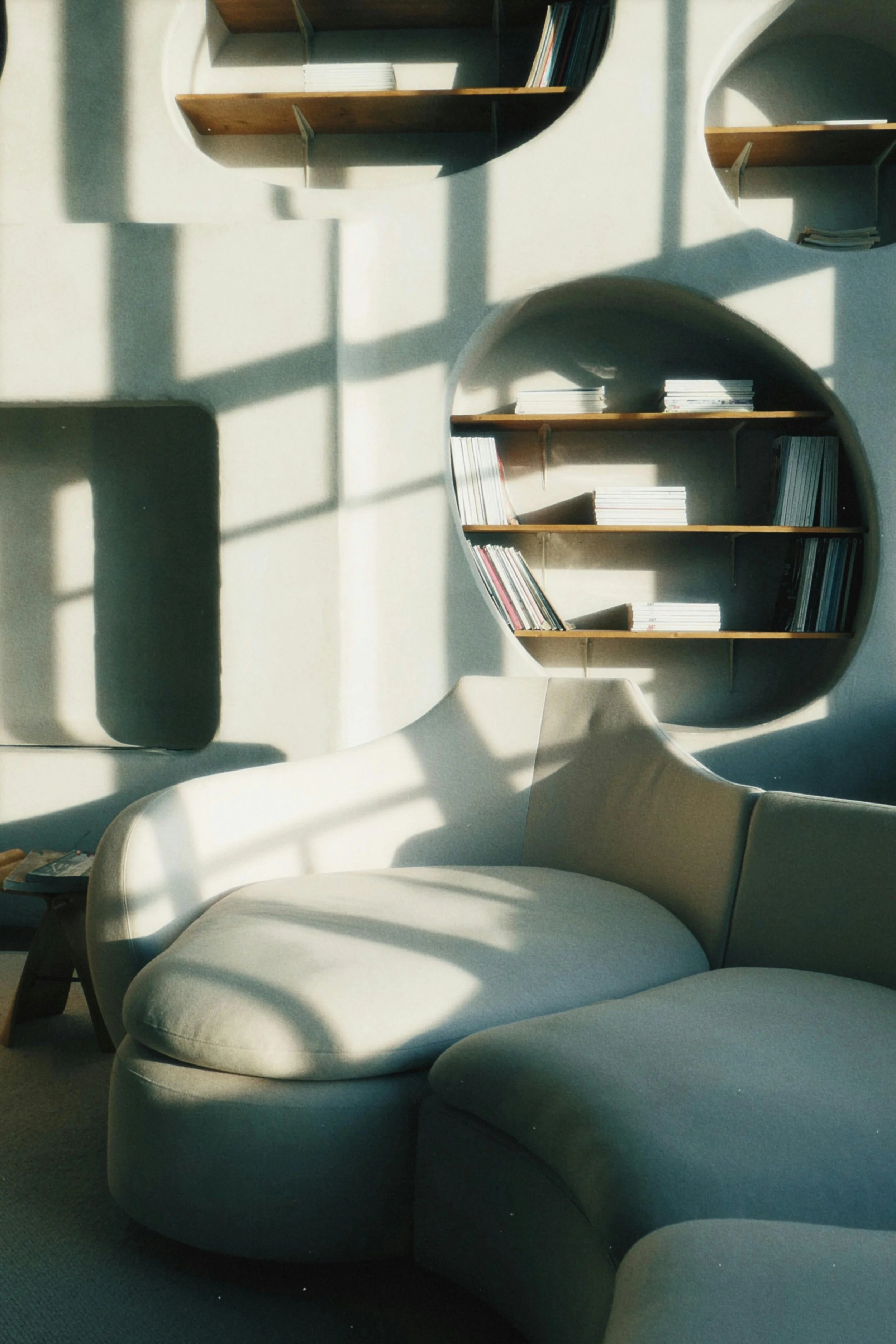 A white armchair with a rounded back and armrest, situated in a room with sunlight casting shadows on the wall. Behind the chair is a decorative wall shelf filled with books, and a small table on the left side holds a magazine.