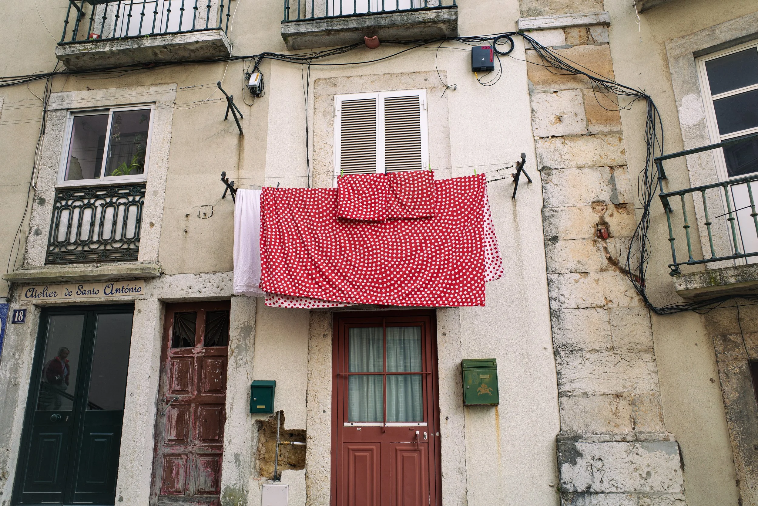 The image shows the facade of an old building with two windows and a door. A clothesline with red and white polka dot fabric is hanging outside, attached to the building. Several wires are running across the facade, and electrical devices are mounted