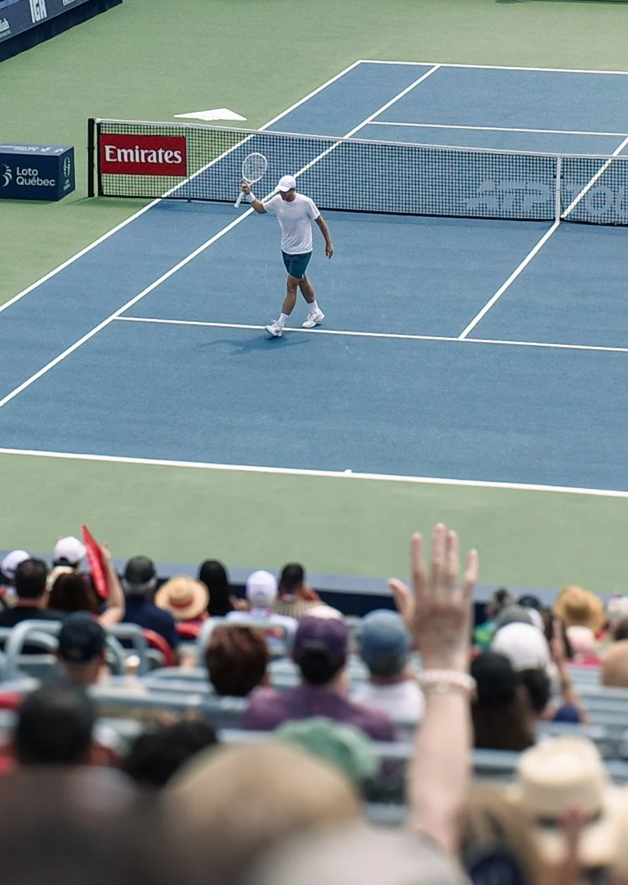 A tennis player on a blue tennis court holding a racket, walking away from the net after a game or practice. Spectators in the stands are watching, with some raising their hands.