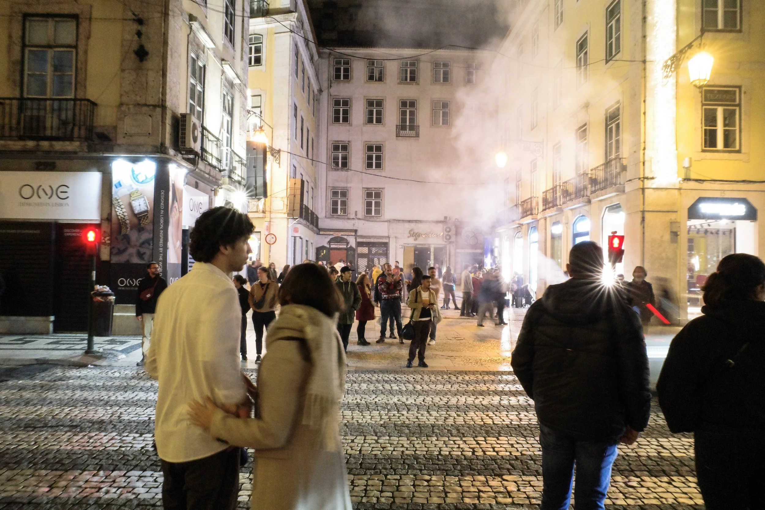 Street scene at night with a crowd of people; buildings are older European style, some smoke or steam rising from the street, illuminated windows and streetlights.