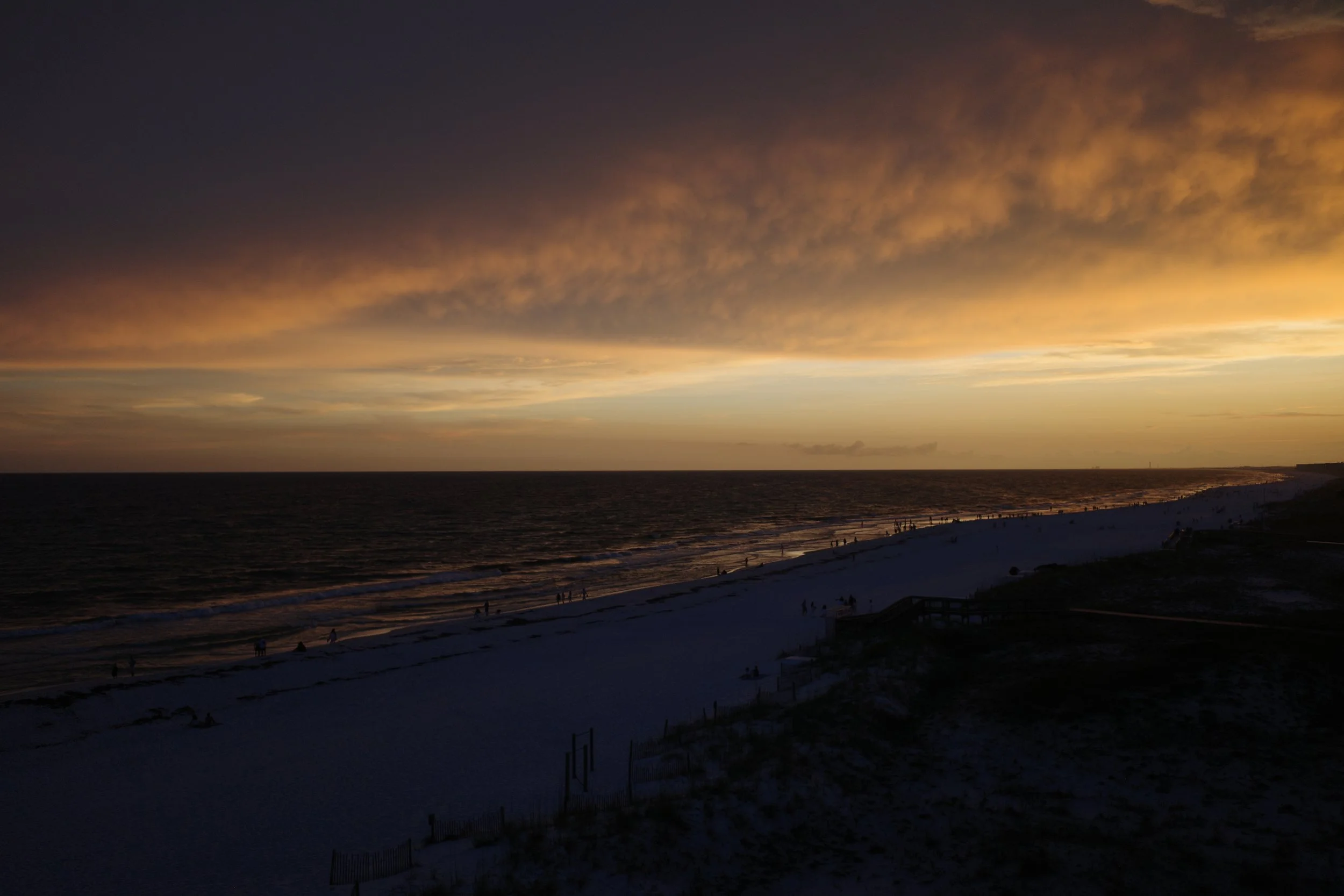 Sunset over the ocean with colorful clouds in the sky and a sandy beach with some people walking along the shoreline.