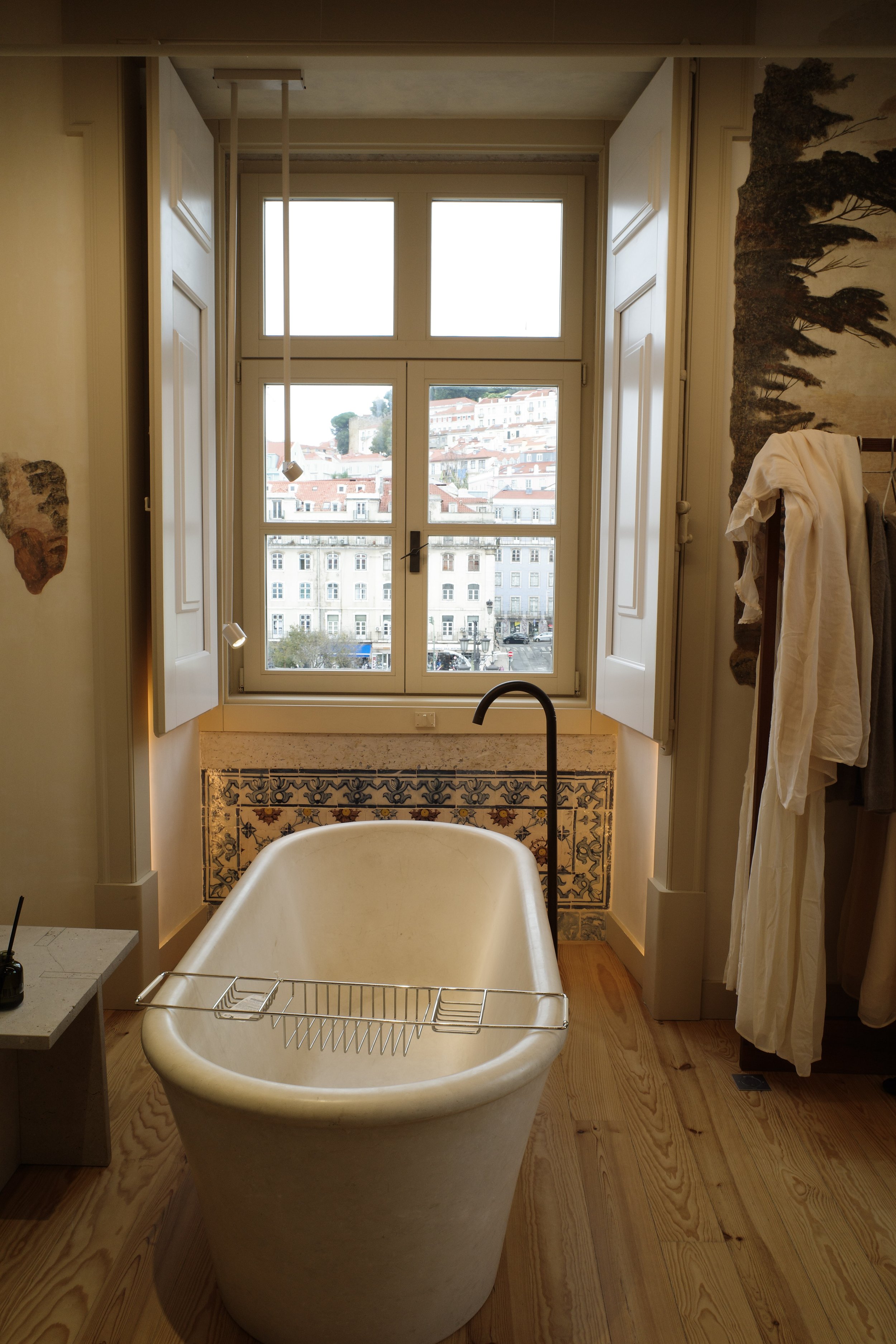 Empty vintage bathtub in a rustic bathroom with wooden flooring, large window with city view, and antique wall decor.