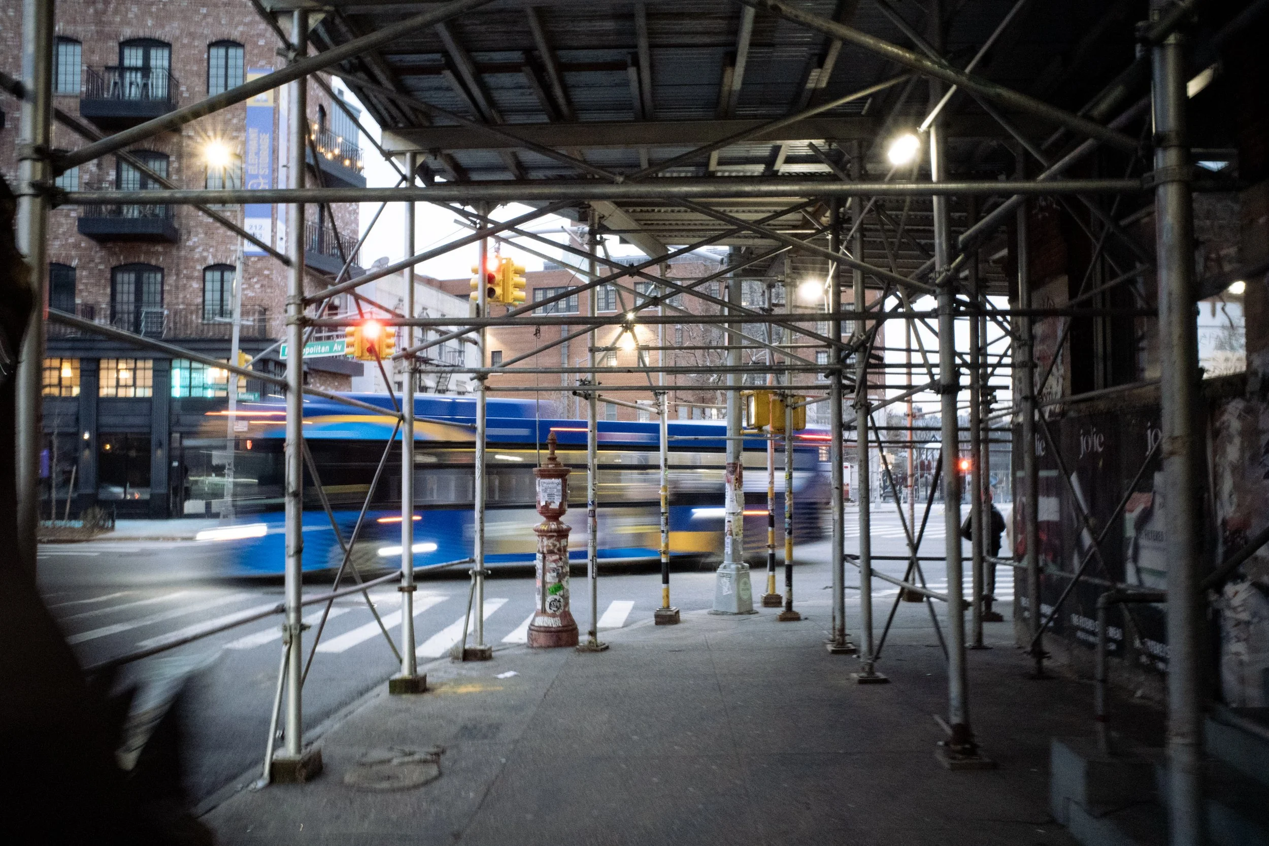 City street scene with scaffolding, a bus passing by, traffic lights, and brick buildings.