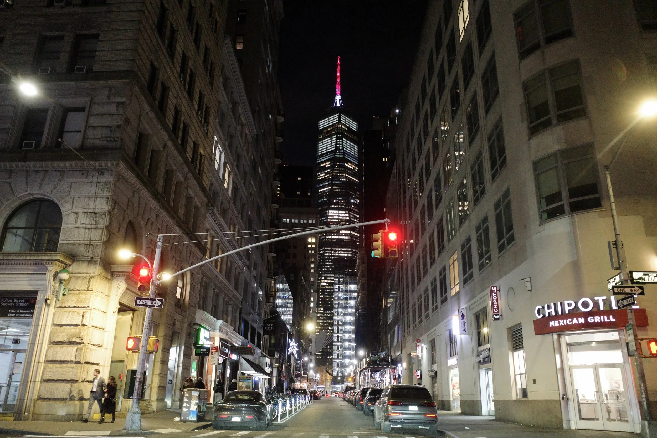 Nighttime street scene with tall buildings, traffic lights, parked cars, pedestrians, and illuminated signs including Chipotle and Subway, with One Way street signs.
