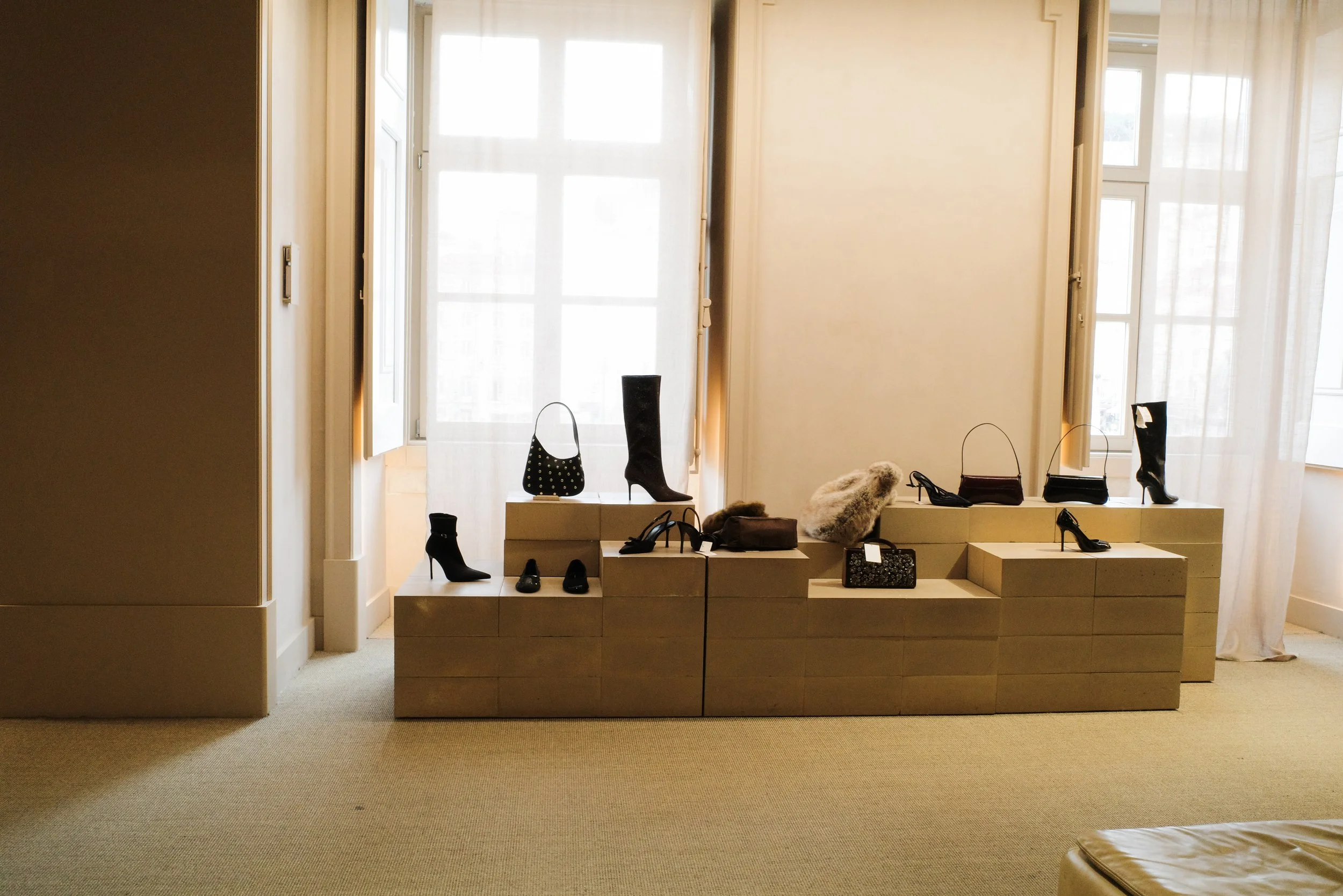 Display of various women's shoes, handbags, and accessories arranged on beige platforms near a large window with sheer curtains in a well-lit room.
