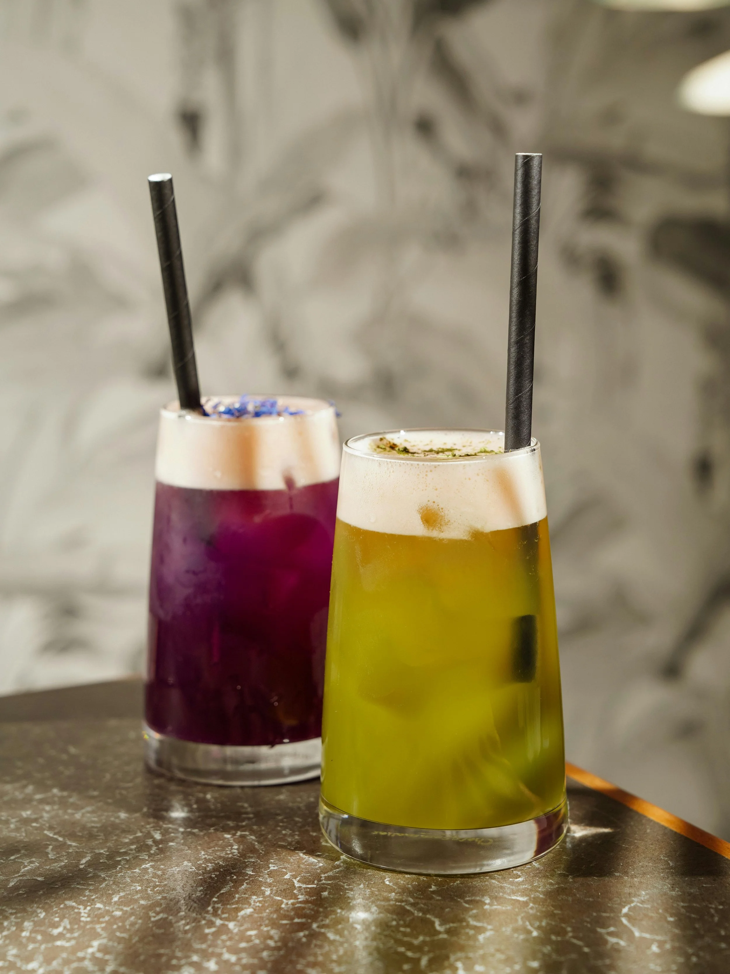 Two colorful beverages in clear glasses with black straws on a textured brown surface, with a blurred background.