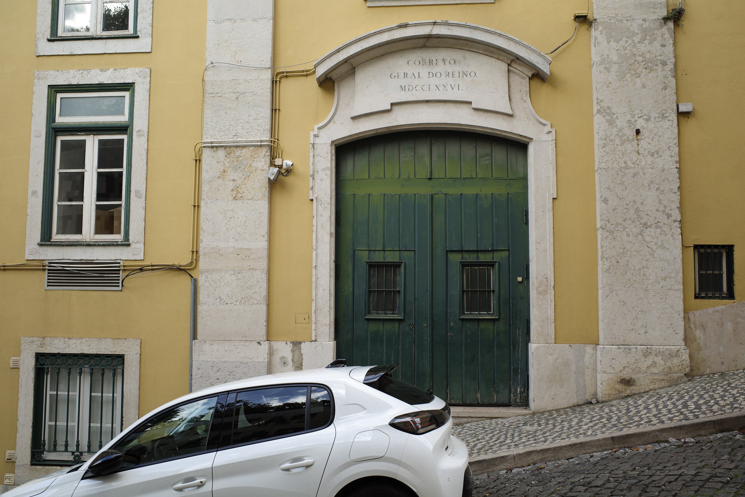 A yellow building with stone accents and a large green arched door, multiple windows with white frames, and a white car parked on a cobblestone street.