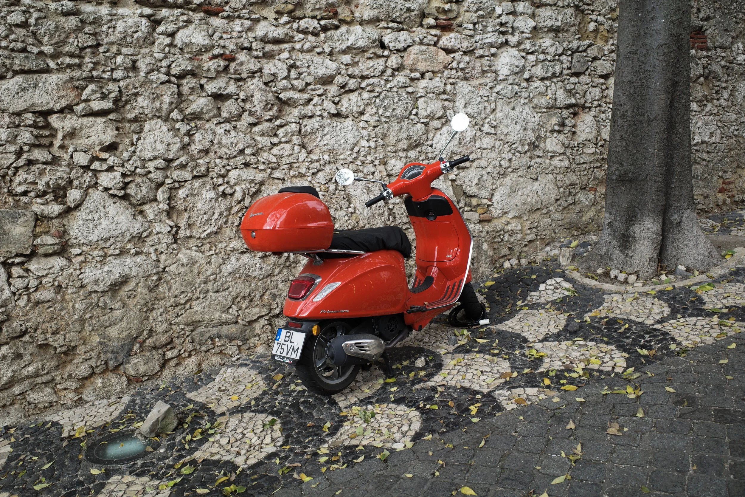 A red scooter parked next to a stone wall and a tree, with a cobblestone and fallen leaves ground.