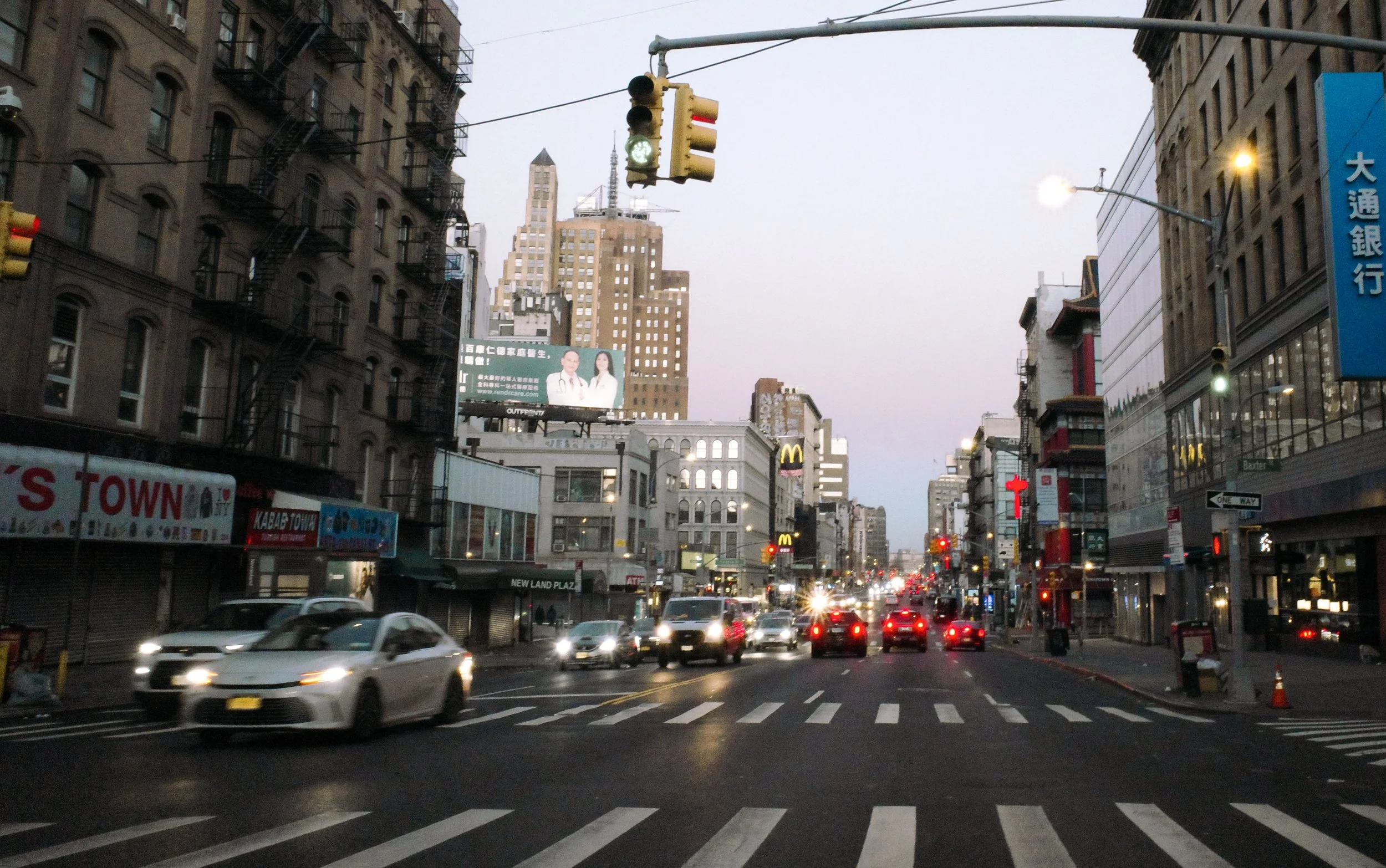 City street scene with traffic lights, vehicles at a busy intersection, tall buildings, and billboards in the background, during early evening or dusk.