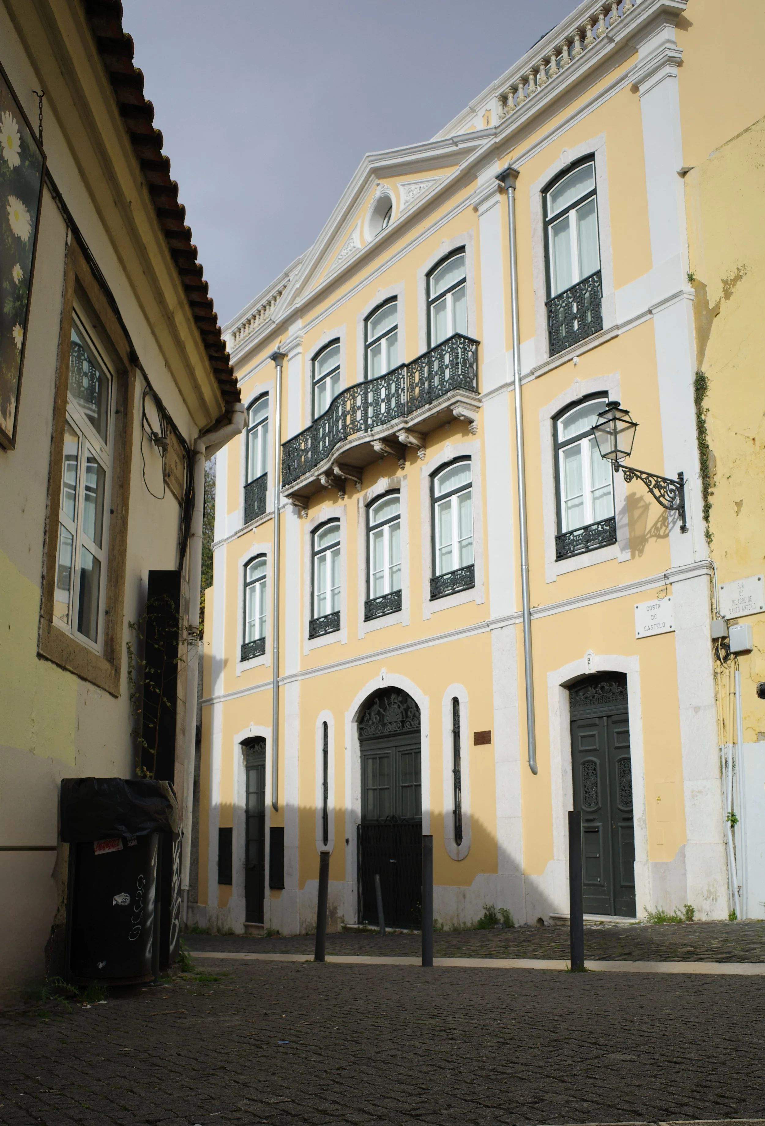 A European-style street scene showing a yellow building with white trim, black wrought-iron balconies, and arched windows. The foreground has a cobblestone street, a black trash bin, and street lamps. The sky is overcast.