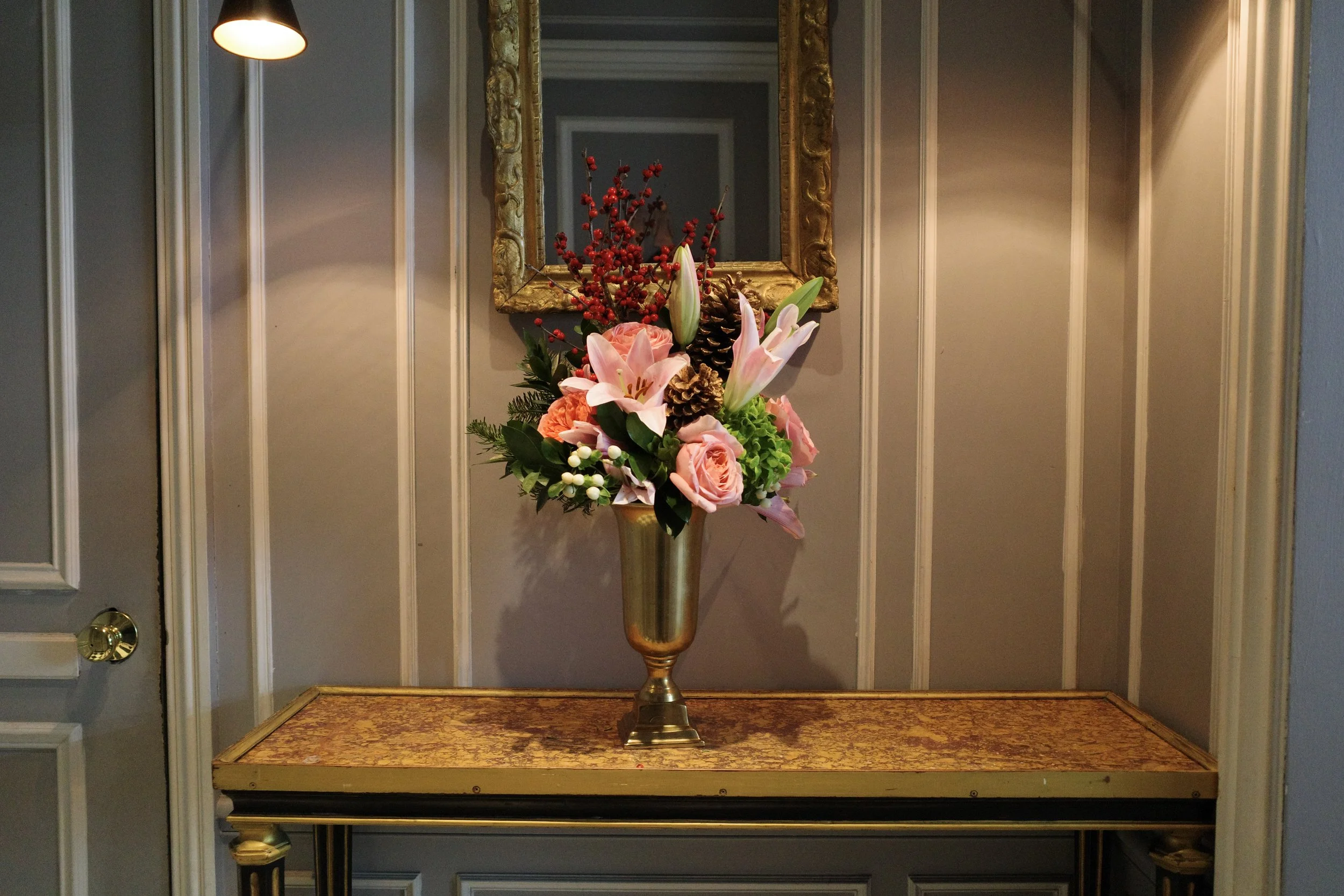 A decorative floral arrangement with lilies, roses, pinecones, and berries in a gold vase, set on a small table in front of a mirror on a wall with paneling.