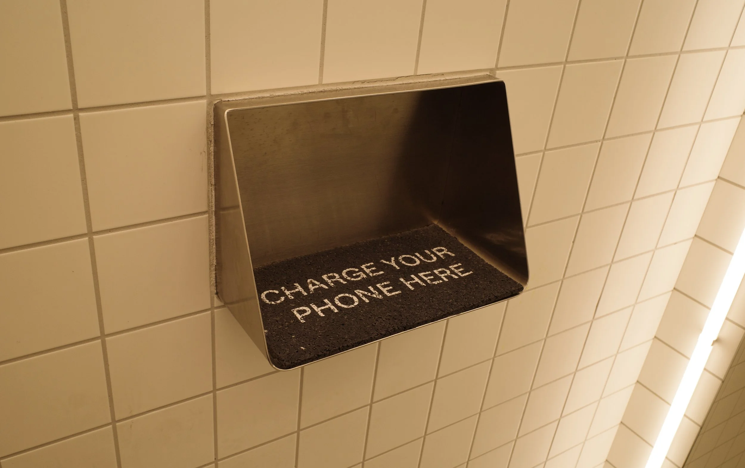 A metal charging station embedded in a tiled wall with a black mat that has white text reading, 'CHARGE YOUR PHONE HERE'.