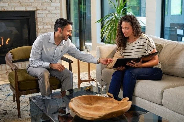 A man in a light gray shirt and gray pants sitting on a vintage armchair, smiling and pointing to a woman's hand. The woman, with curly hair, wearing a striped shirt and dark pants, seated on a beige sofa holding a black folder or tablet. They are in a modern living room with a glass coffee table, a wooden bowl, a glass of water, a fireplace, and large windows with plants outside.