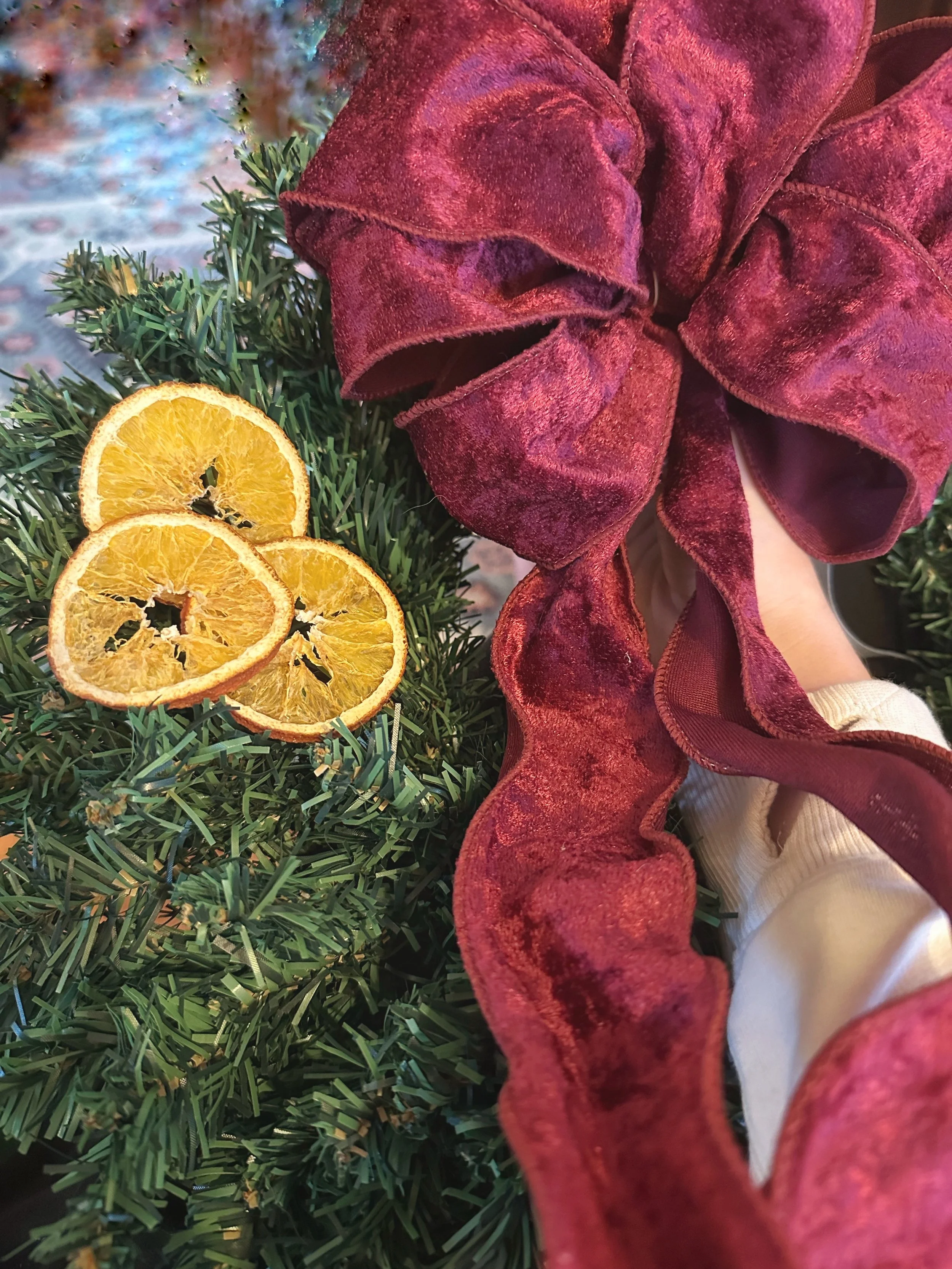 Close-up of dried orange slices at the base of a Christmas arrangement, with a red velvet ribbon and green artificial pine branches in the background.