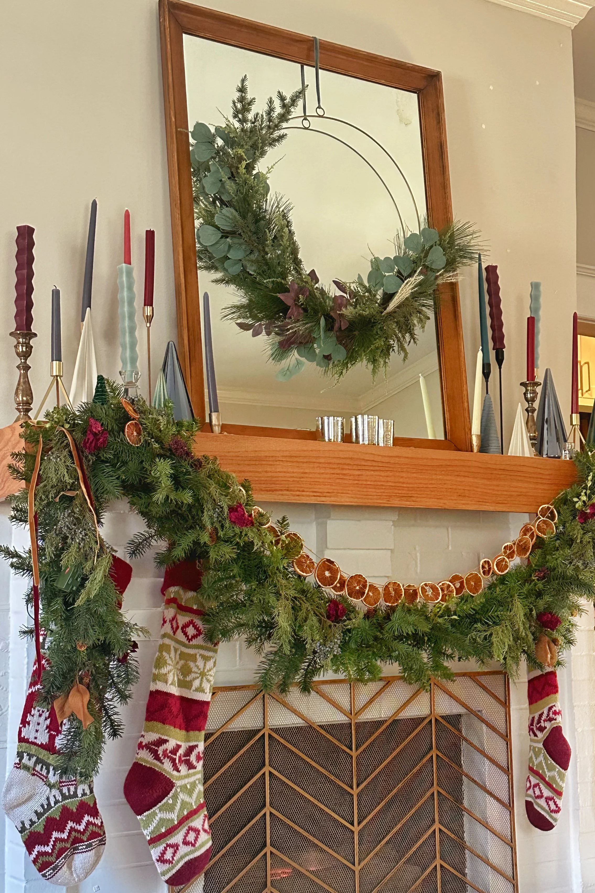 Decorated fireplace mantel with Christmas stockings, greenery, and a mirror with a hanging wreath reflected in it.