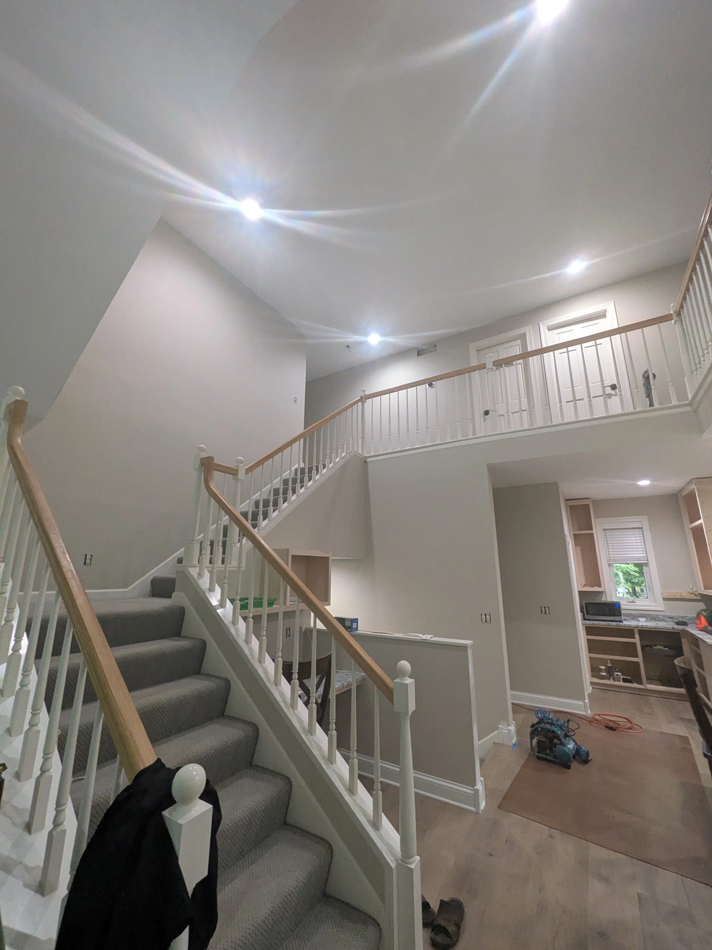 Interior of a house under construction or renovation, featuring a staircase with gray carpeted steps and a wooden handrail, an open kitchen area with partially installed cabinets, and construction tools and materials on the floor.