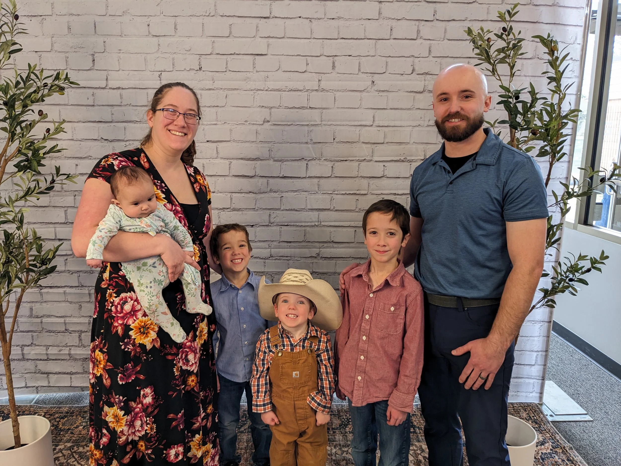 A smiling woman holding a baby girl in a floral dress stands next to a man with a beard, surrounded by four children, in front of a white brick wall with potted trees on either side.