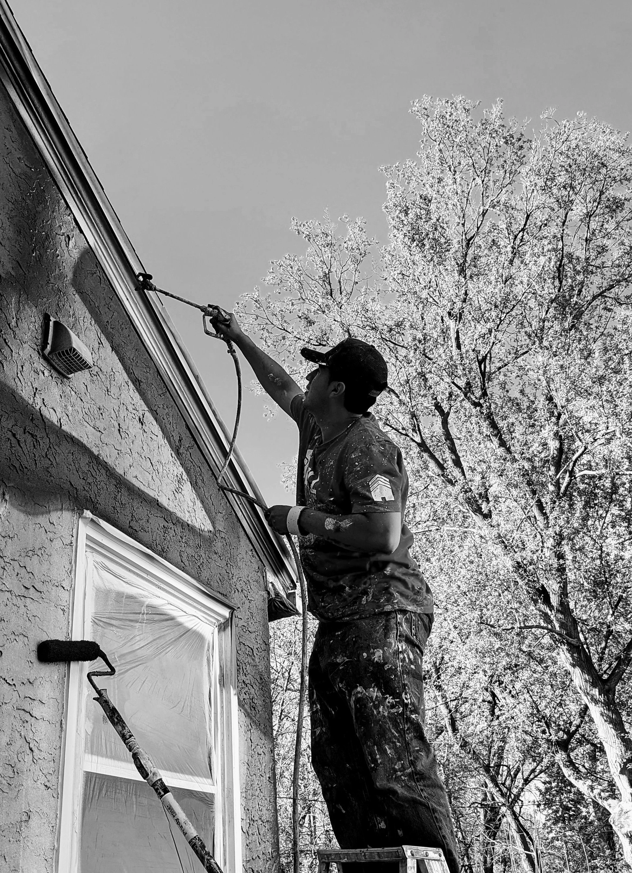 A person wearing a baseball cap and camouflage pants is on a ladder cleaning or inspecting the exterior of a house's window with a spray or cleaning tool. Trees with bare branches are in the background.