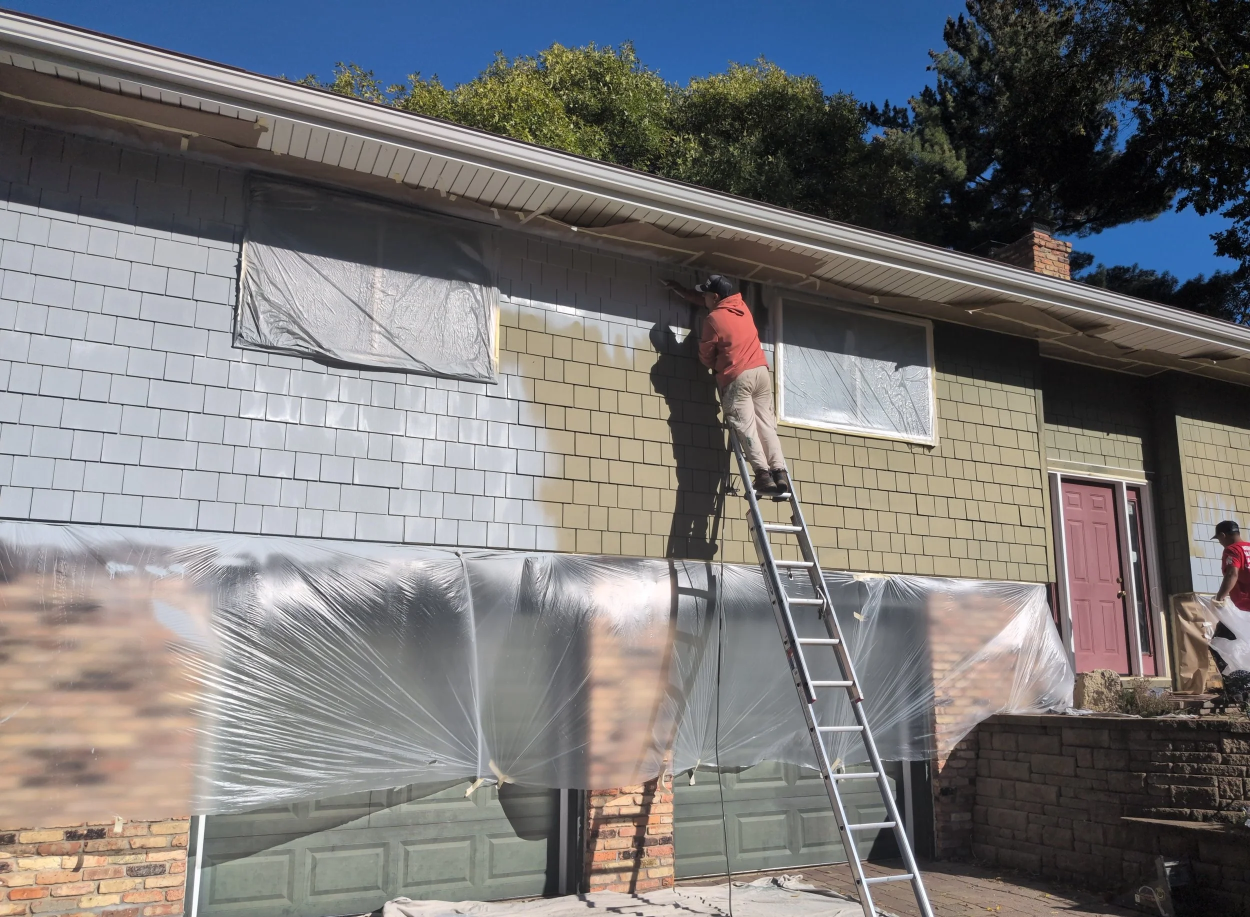 Person painting the exterior wall of a house while standing on a ladder, with windows covered in plastic for protection, and trees in the background on a clear, sunny day.
