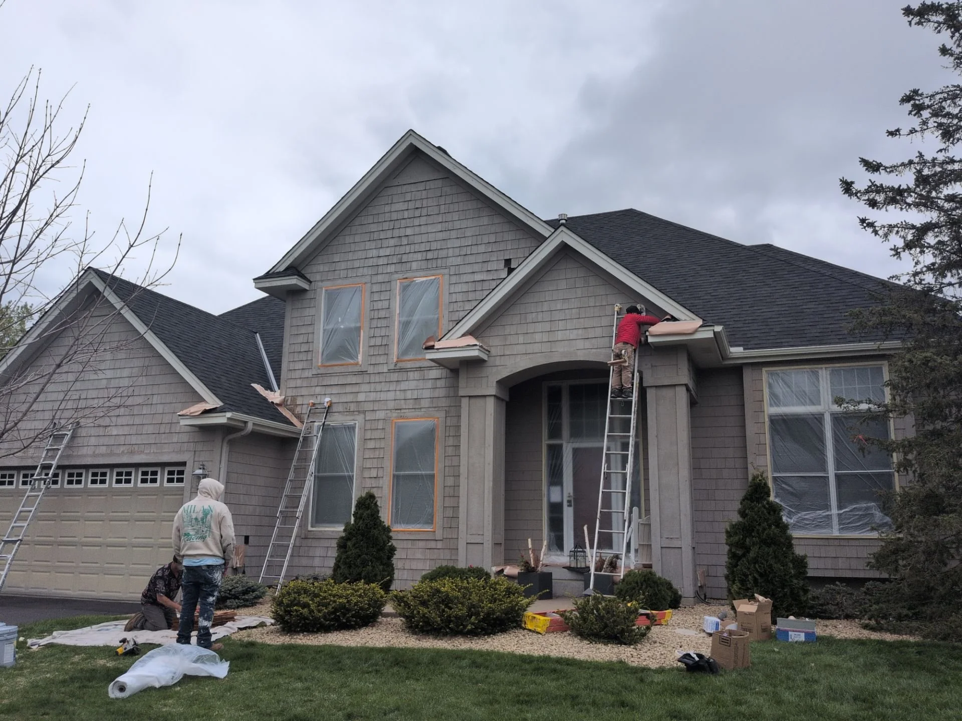 Homeowner applying new roof shingles on the roof of a two-story house, with contractors supervising and working on the edge, and tools and supplies scattered on the lawn in front.