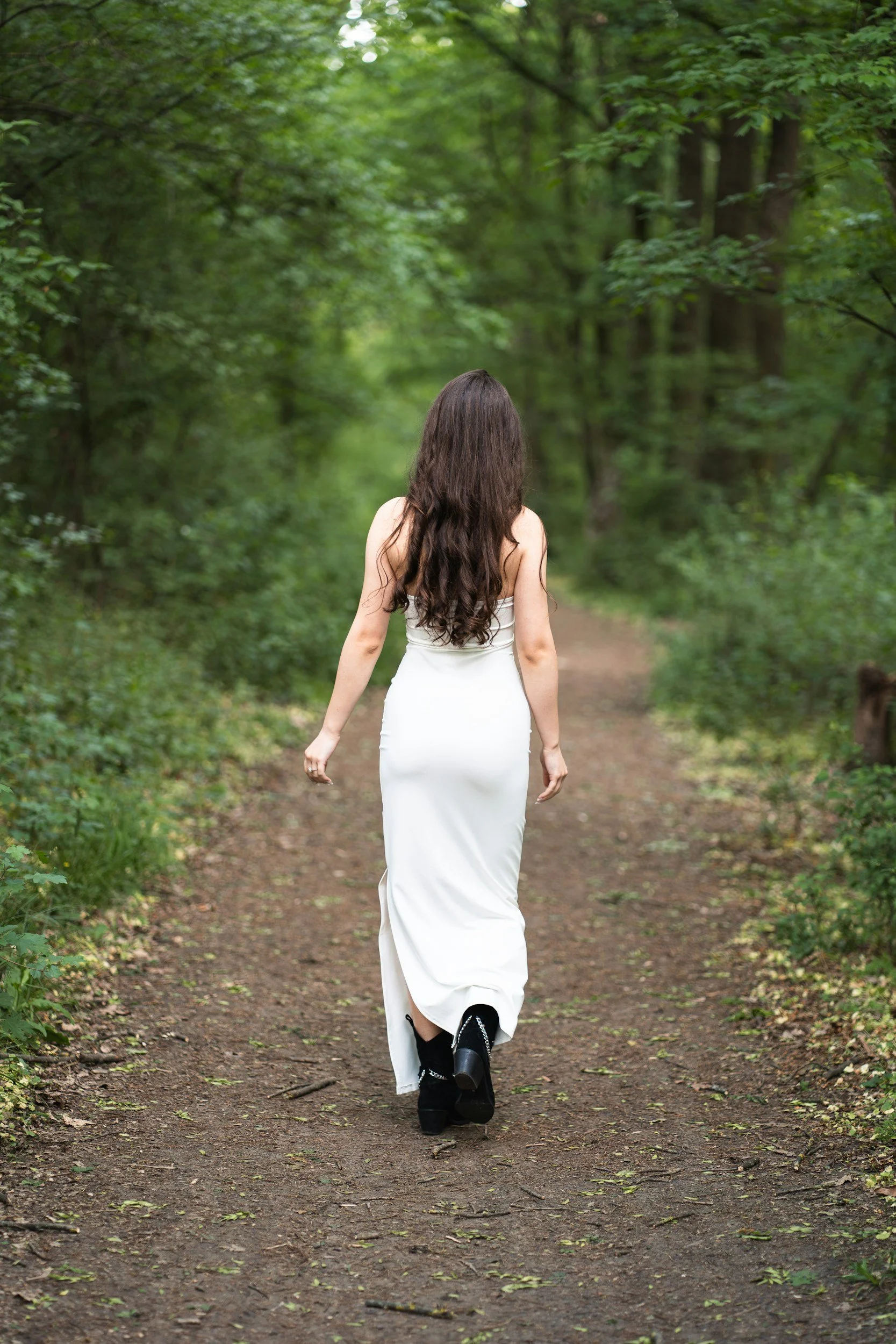 A woman with long brown hair walking down a forest trail surrounded by green trees, wearing a white dress and black boots.