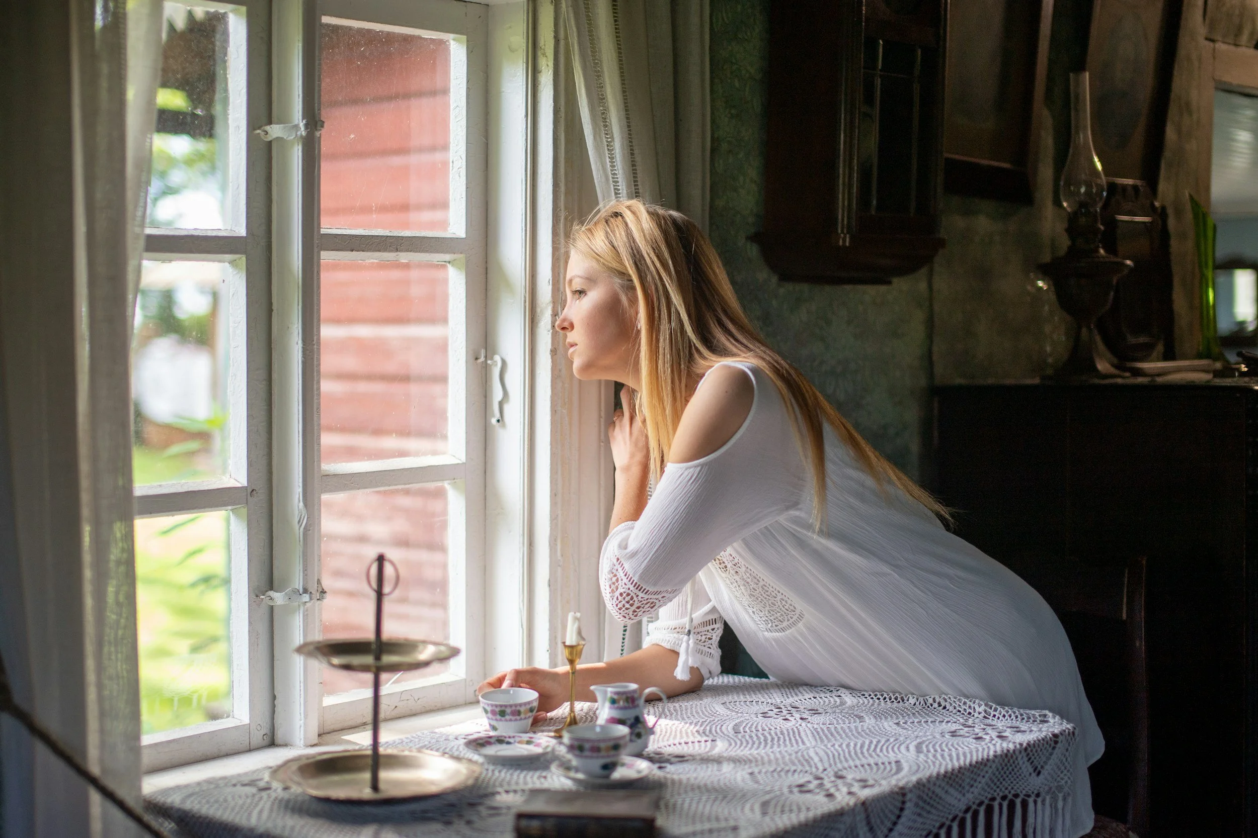 A woman with red hair wearing a white dress gazes out a window splattered with rain, standing next to a table with tea cups and a lace tablecloth in a rustic kitchen.