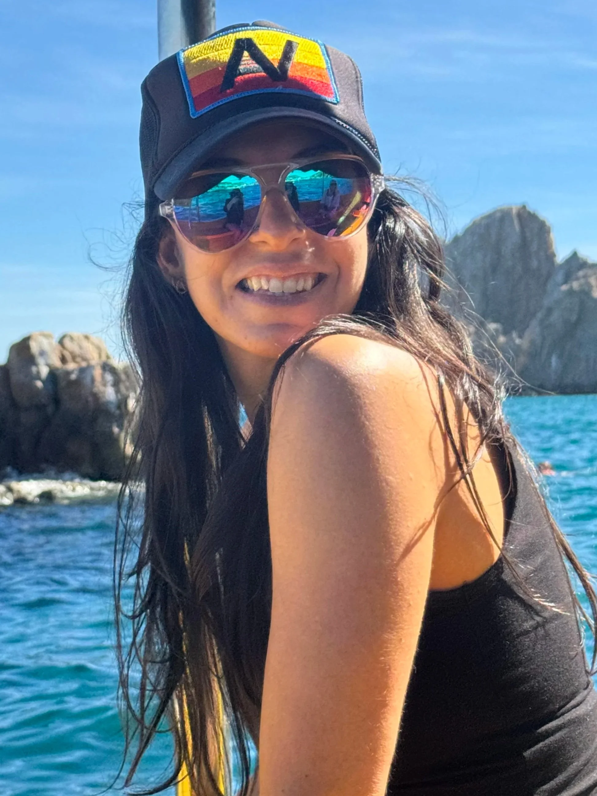 A woman with long dark hair wearing sunglasses and a cap with an 'N' patch, smiling, in front of rocky cliffs and the ocean on a sunny day.