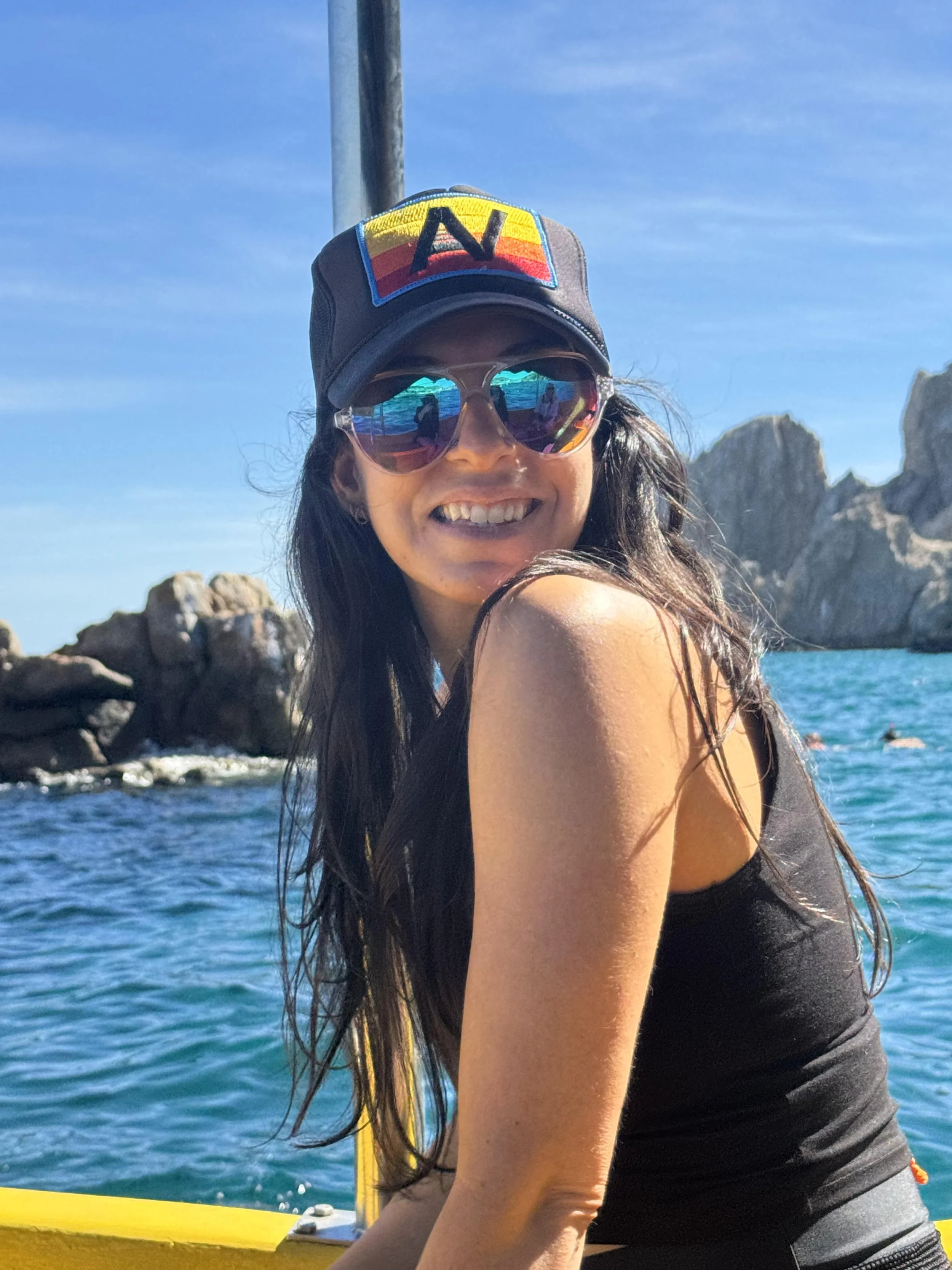 A woman with long dark hair, wearing a black cap with a colorful patch and sunglasses, smiling while on a boat in a blue ocean with rocky islands in the background.
