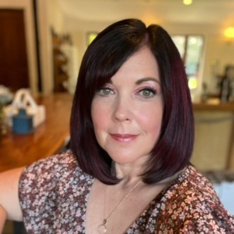 A woman with shoulder-length dark hair and light eyes takes a selfie in a cozy, well-lit home interior. She is wearing a floral top and a necklace.