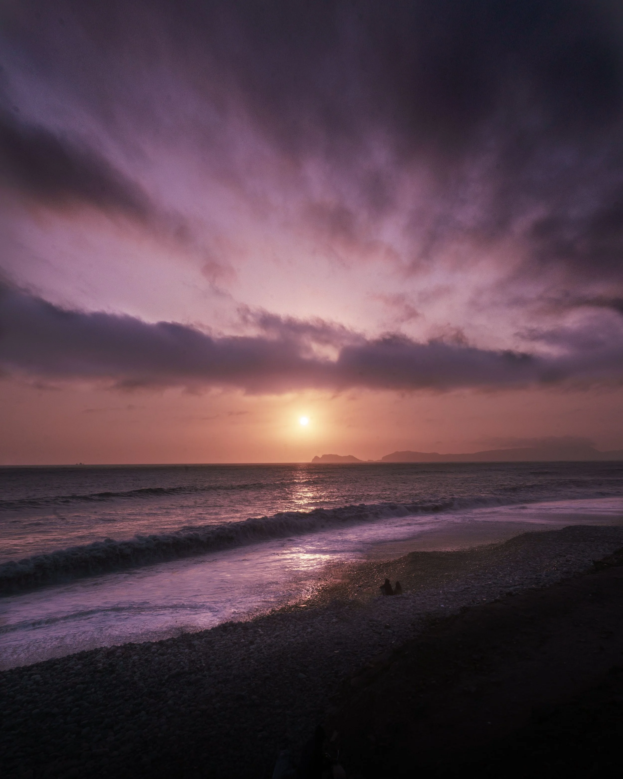 Sunset over the ocean with dark clouds and a rocky beach in the foreground.