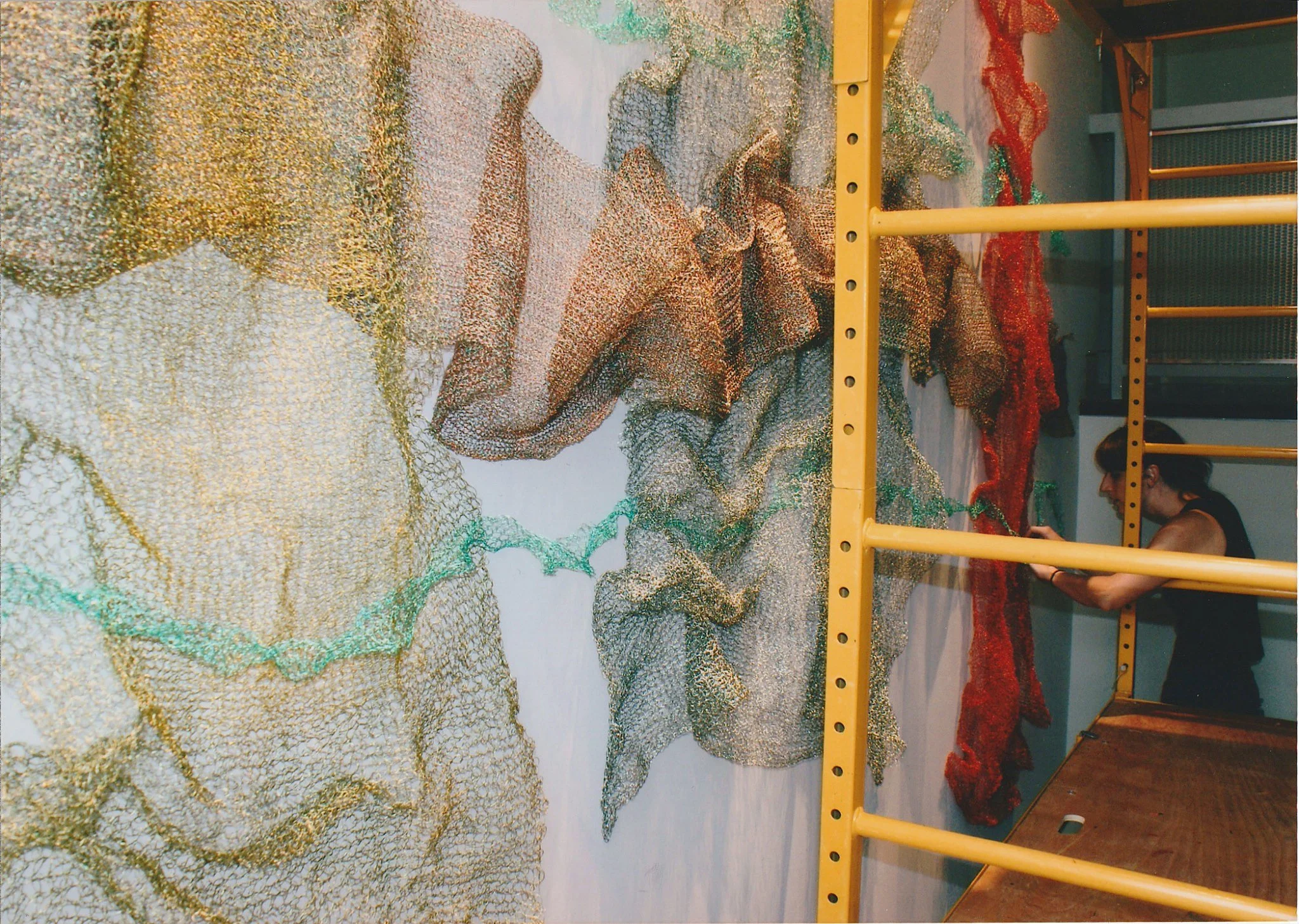 Colorful textured artwork in an indoor space with people sitting near it, yellow metal structure in front of the artwork.