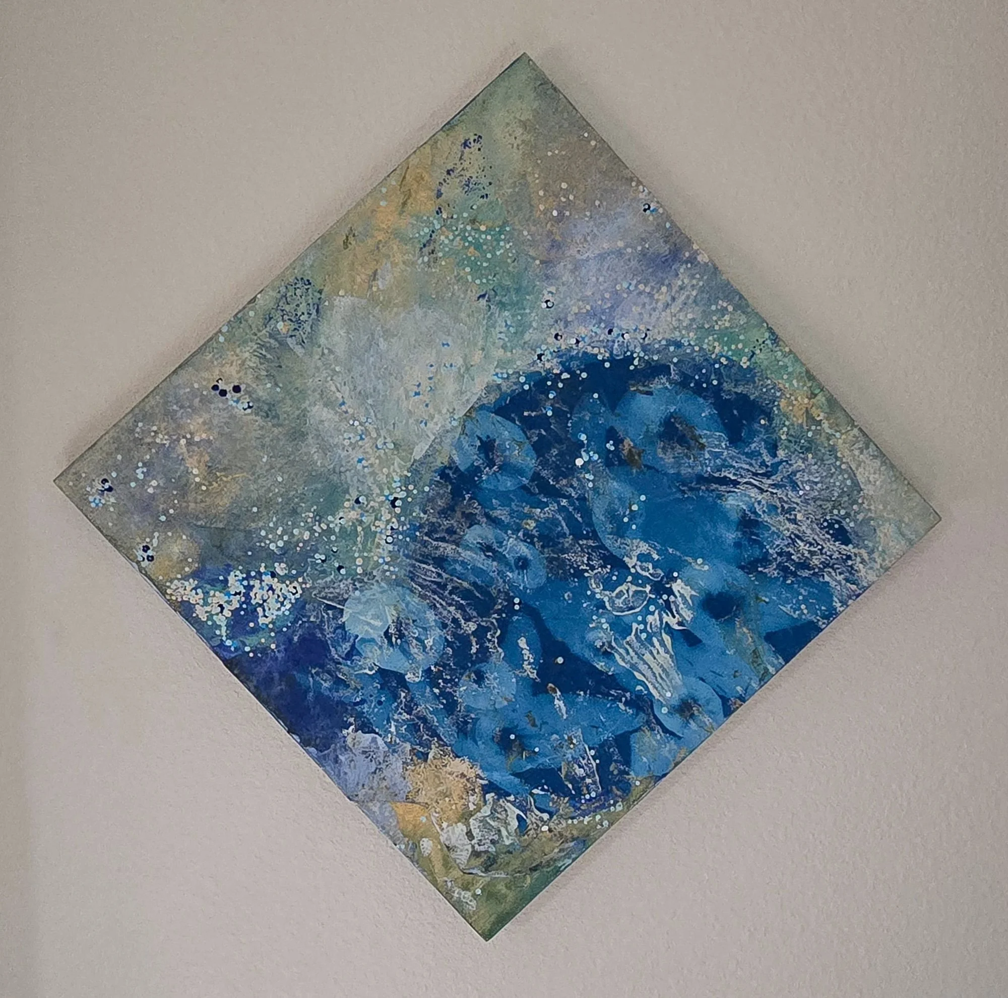 An abstract watercolor painting with shades of blue, green, and hints of yellow, arranged in a geometric diamond shape on a white surface.