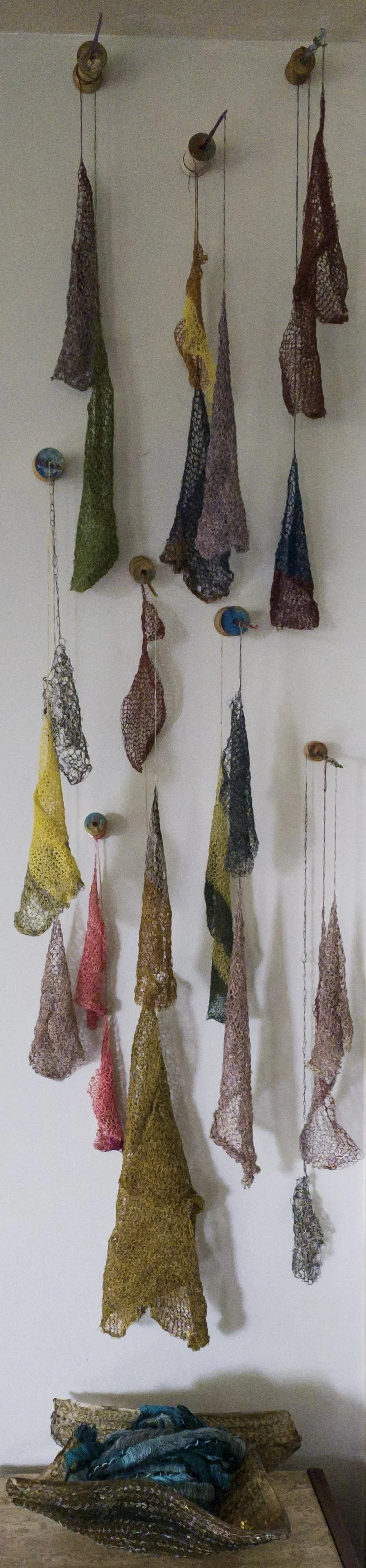 Multiple colorful crocheted scarves hanging from wooden hooks on a white wall, with some scarves stored in a wooden bowl on the floor.