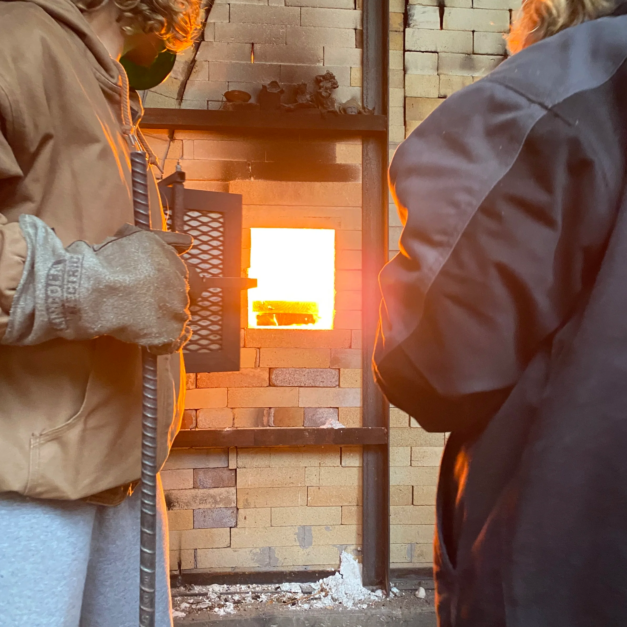 Two people sitting in front of a fireplace made of brick, with fire burning. They are seen from the side and partially in the frame, with only their shoulders and heads visible.