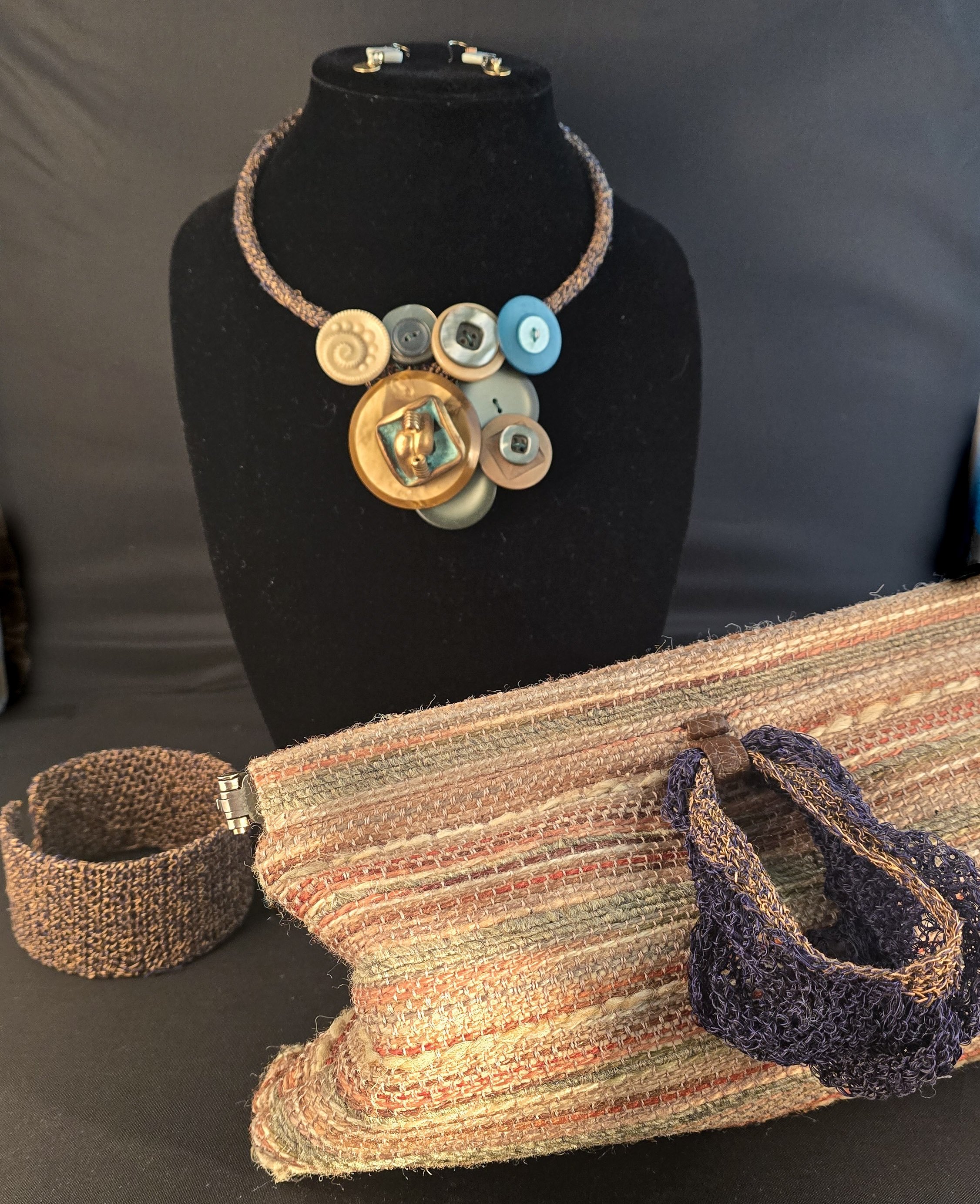 Display of fashion accessories including a woven straw hat with a multicolored pattern, a woven purse with a colorful design and navy blue drawstrings, and a black velvet bust with a chunky statement necklace made of large buttons and shells, and matching earrings.