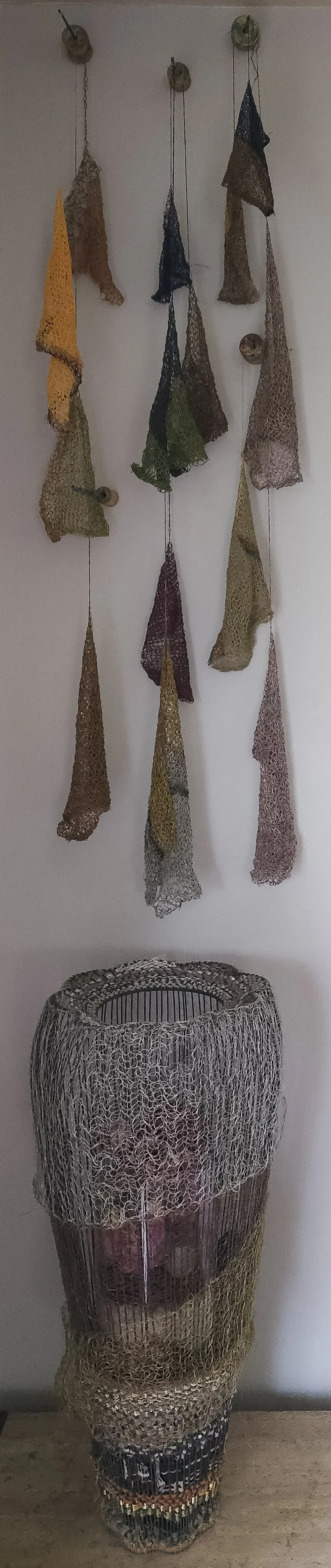 Colorful crocheted and knitted scarves hanging on hooks above a wire sculpture of a human face.
