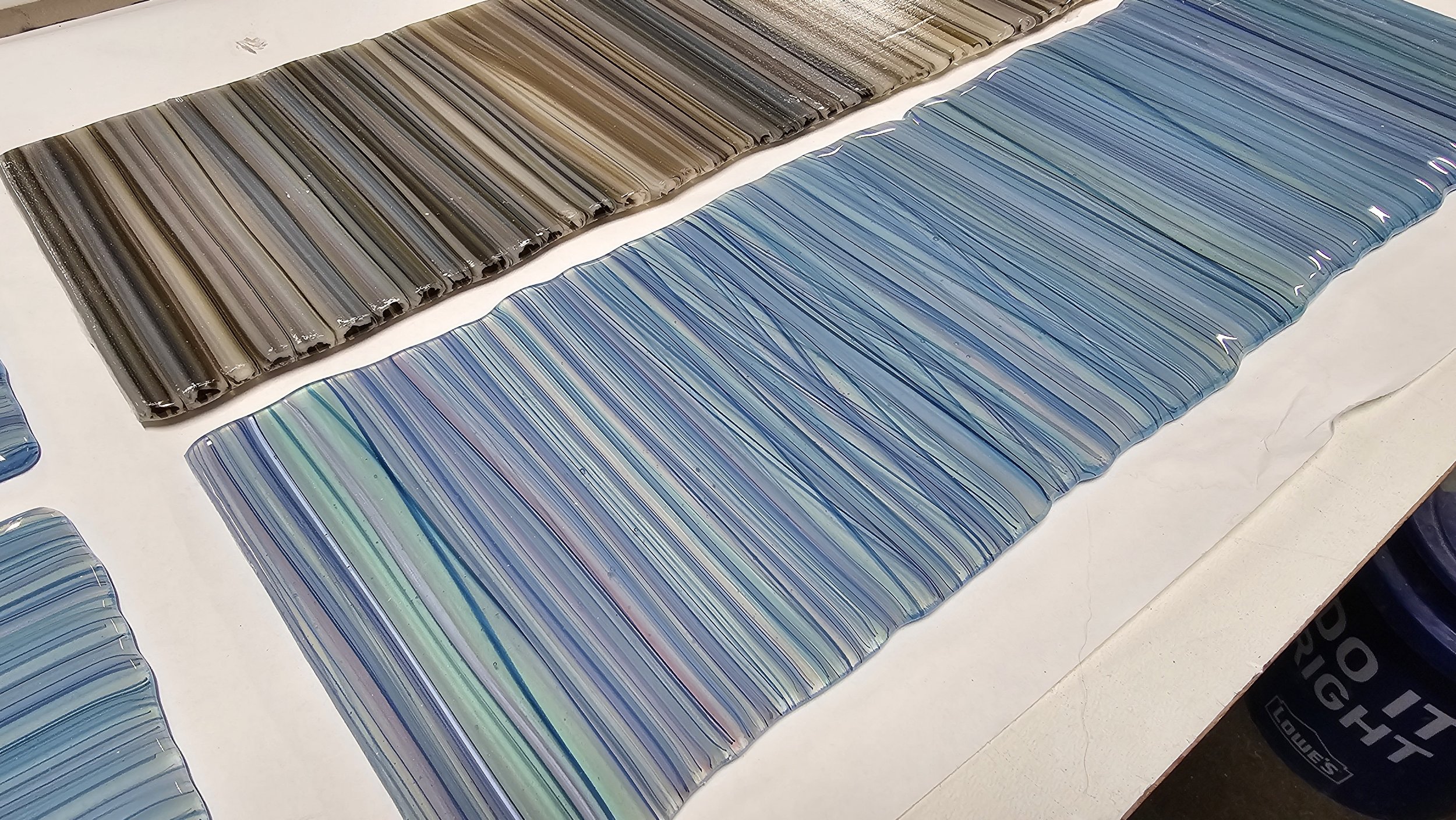 Sheets of glass with colored stripes in various shades of brown, blue, and iridescent hues, laid out on a white surface.