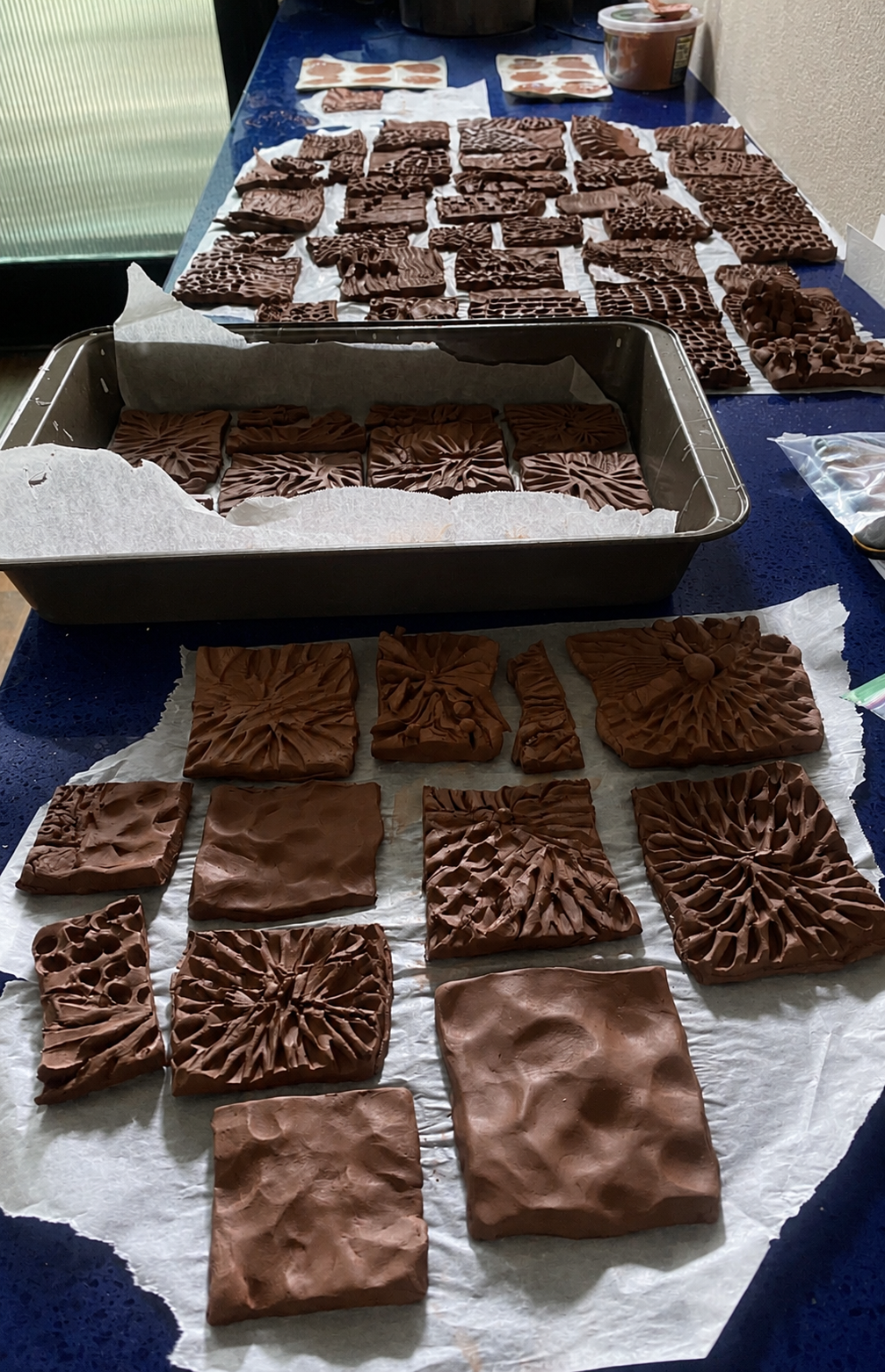 Chocolate soap bars with various textured patterns laid out on a blue table and a baking sheet, with a container of chocolate and some wrapped items in the background.