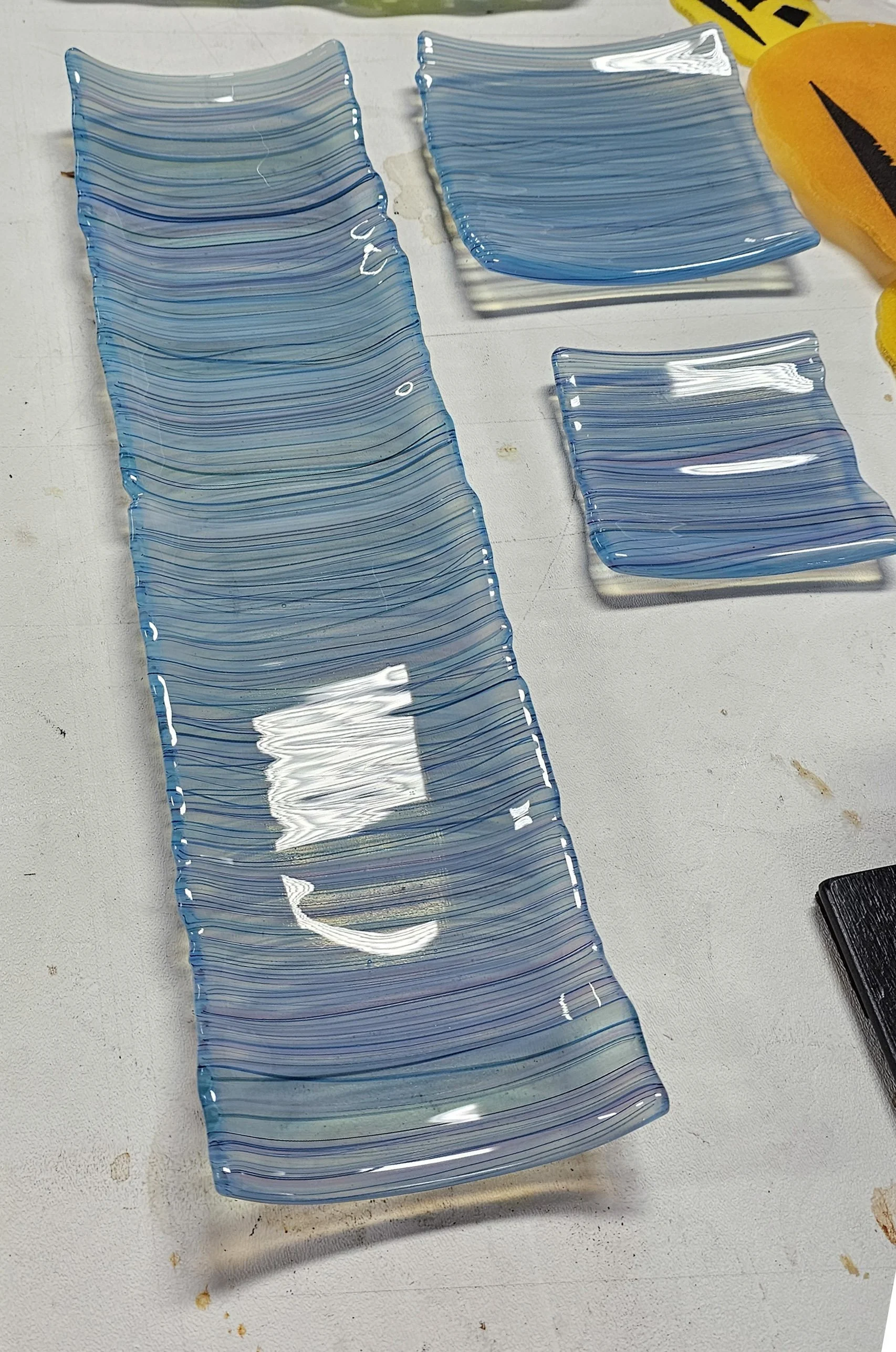 Several glass art pieces with blue and clear horizontal lines arranged on a white table, partially visible yellow object on the right side.