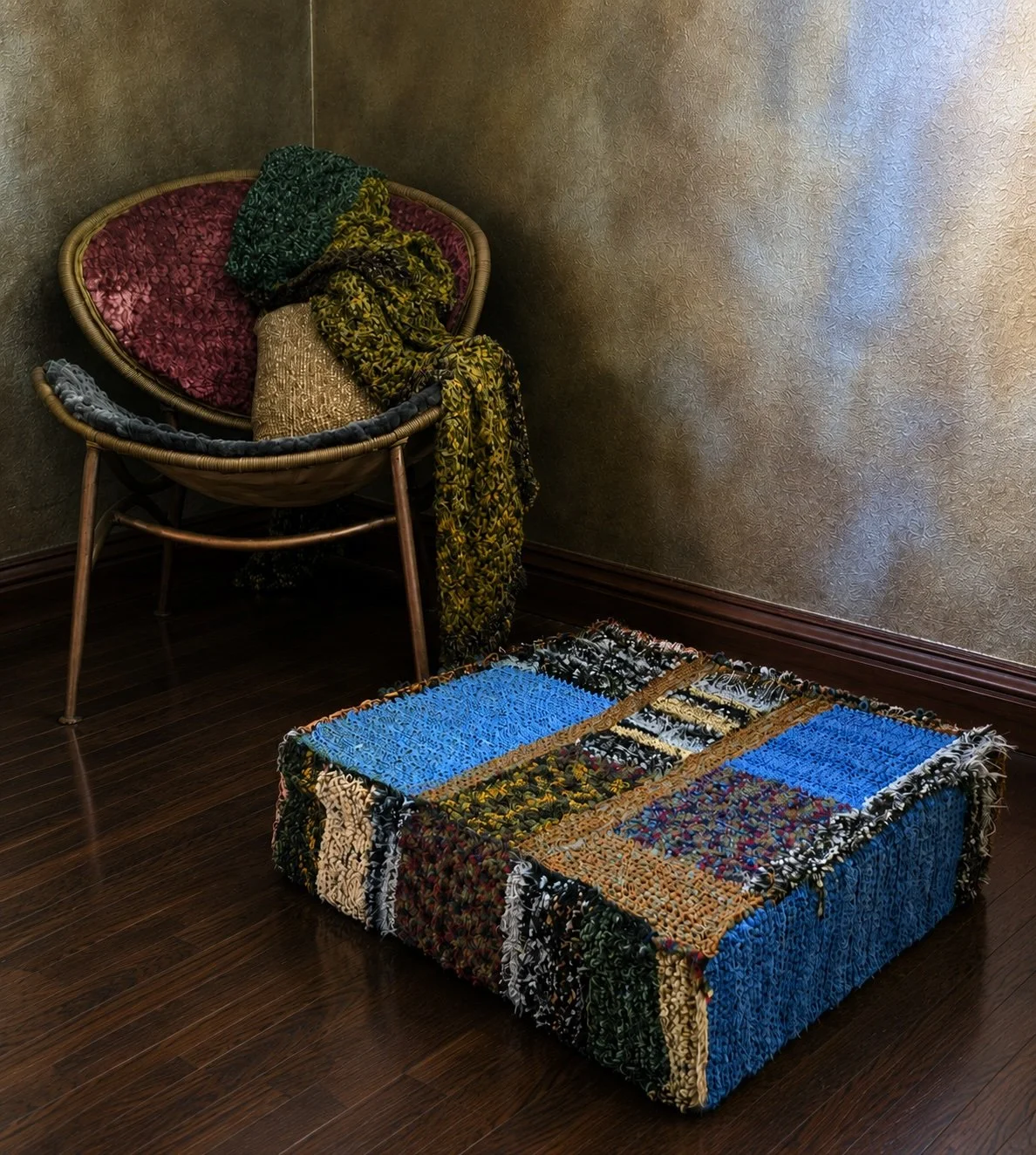 A woven chair with a pink cushion, draped with patterned scarves in green, yellow, and black. In front, a box is covered with a patchwork quilt in various colors, sitting on dark wooden floor, against a textured beige wall.