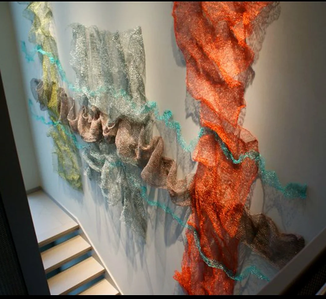 Colorful abstract fabric wall art with flowing, textured layers in shades of silver, brown, orange, and blue, hanging on a white wall.