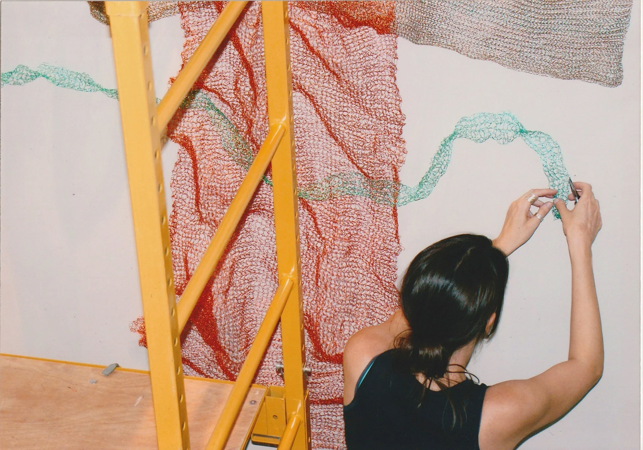 Person working on a large, colorful wire art piece on a white wall, with a yellow leaning ladder in the foreground.