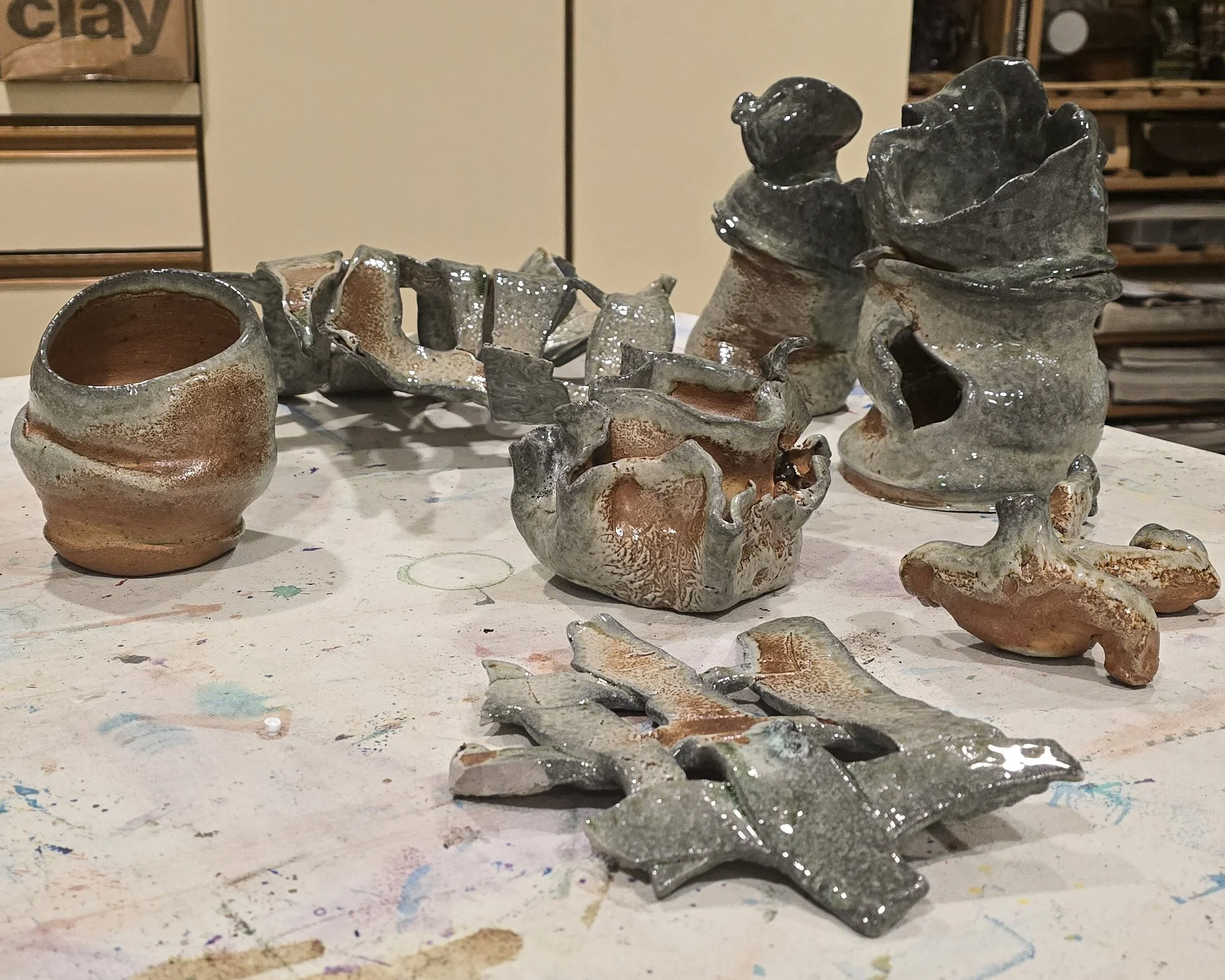 Collection of partially glazed ceramic pottery and sculptures on a paint-stained surface.