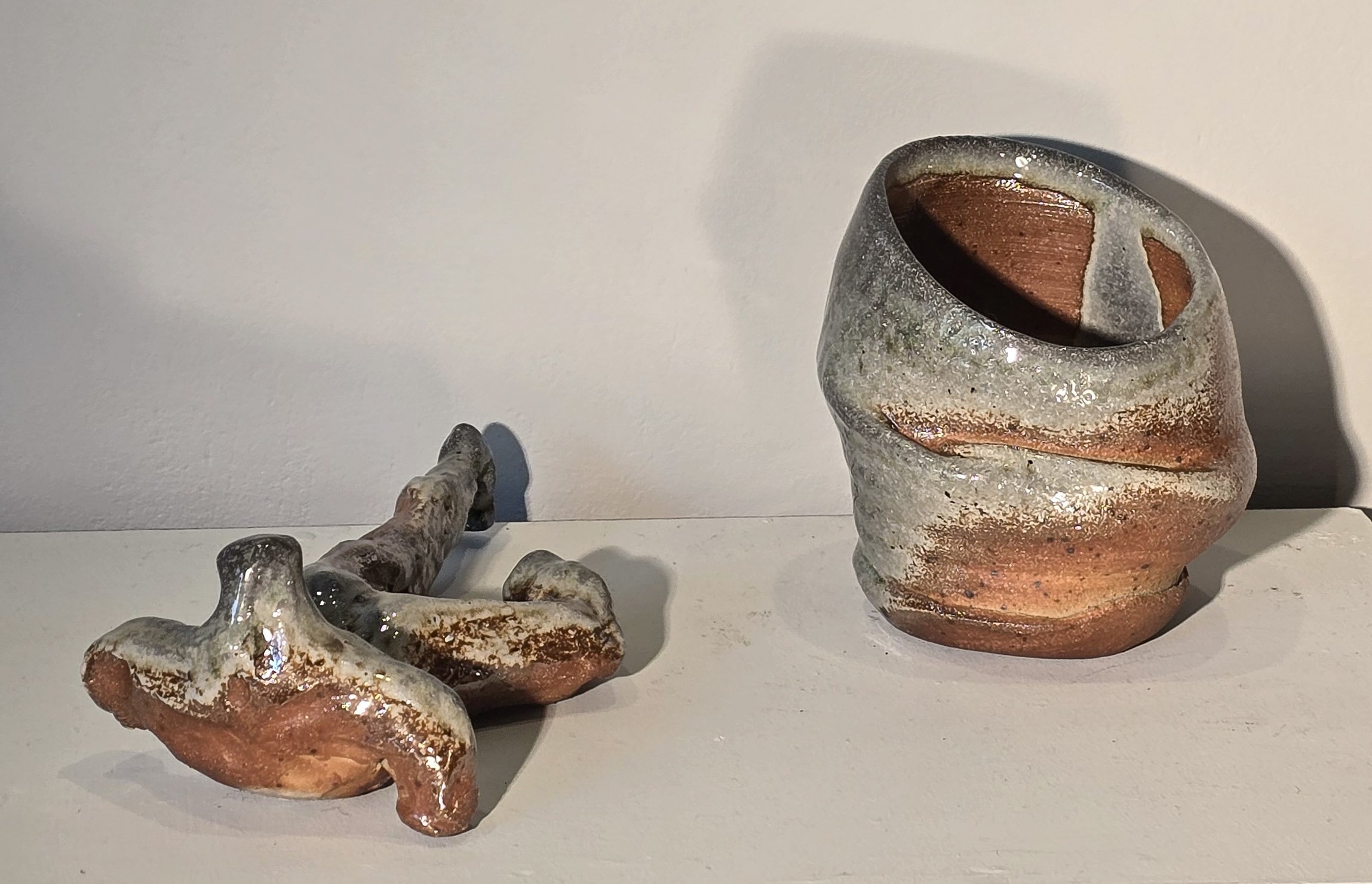 Ceramic or clay sculptures of abstract shapes and a clay vessel with a textured surface, placed on a white surface against a white wall.