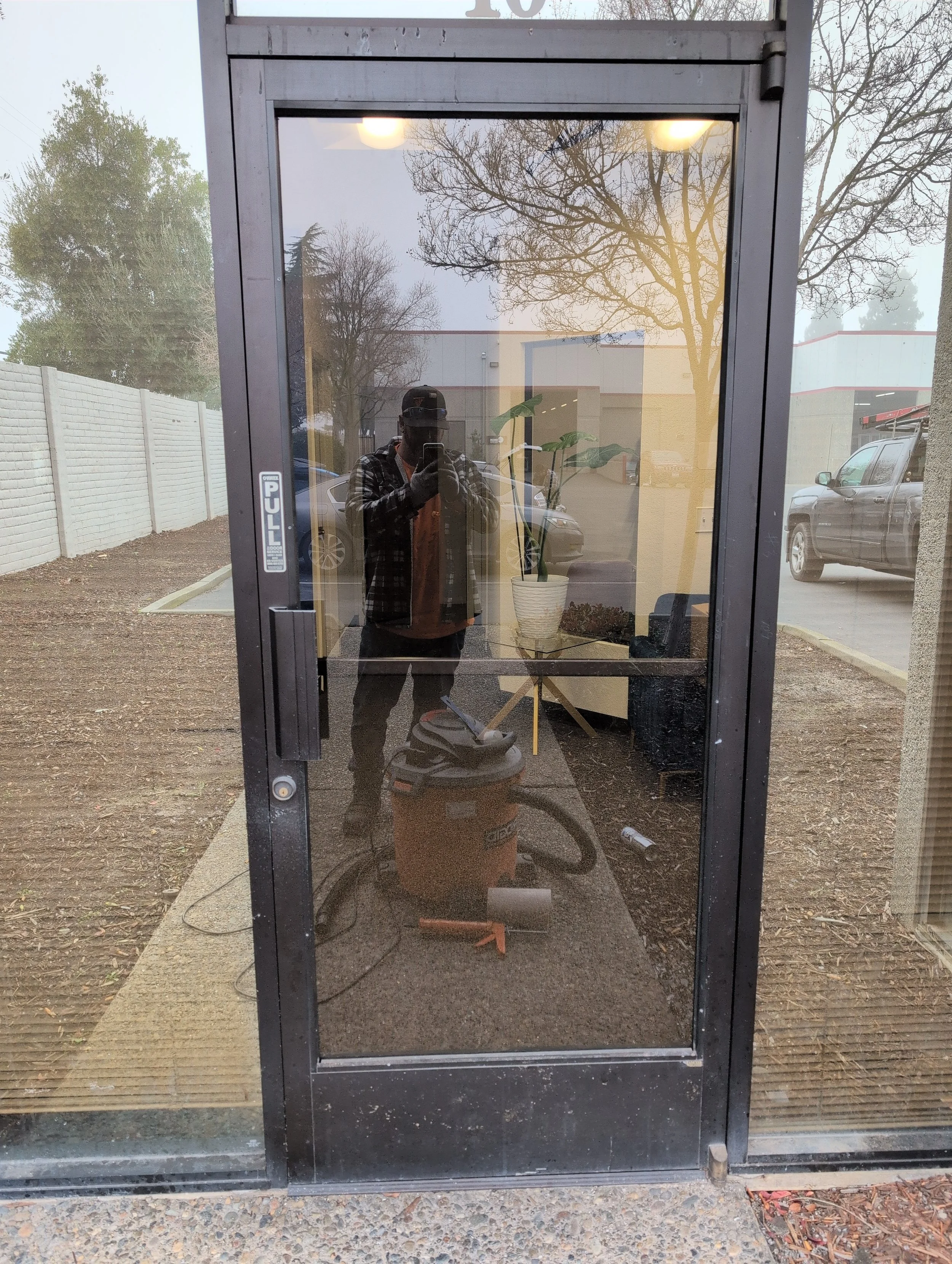 Reflected image of a man wearing a black cap and checkered jacket, taking a photo with a cellphone inside a glass door, with a plant, parked cars, and trees outside.