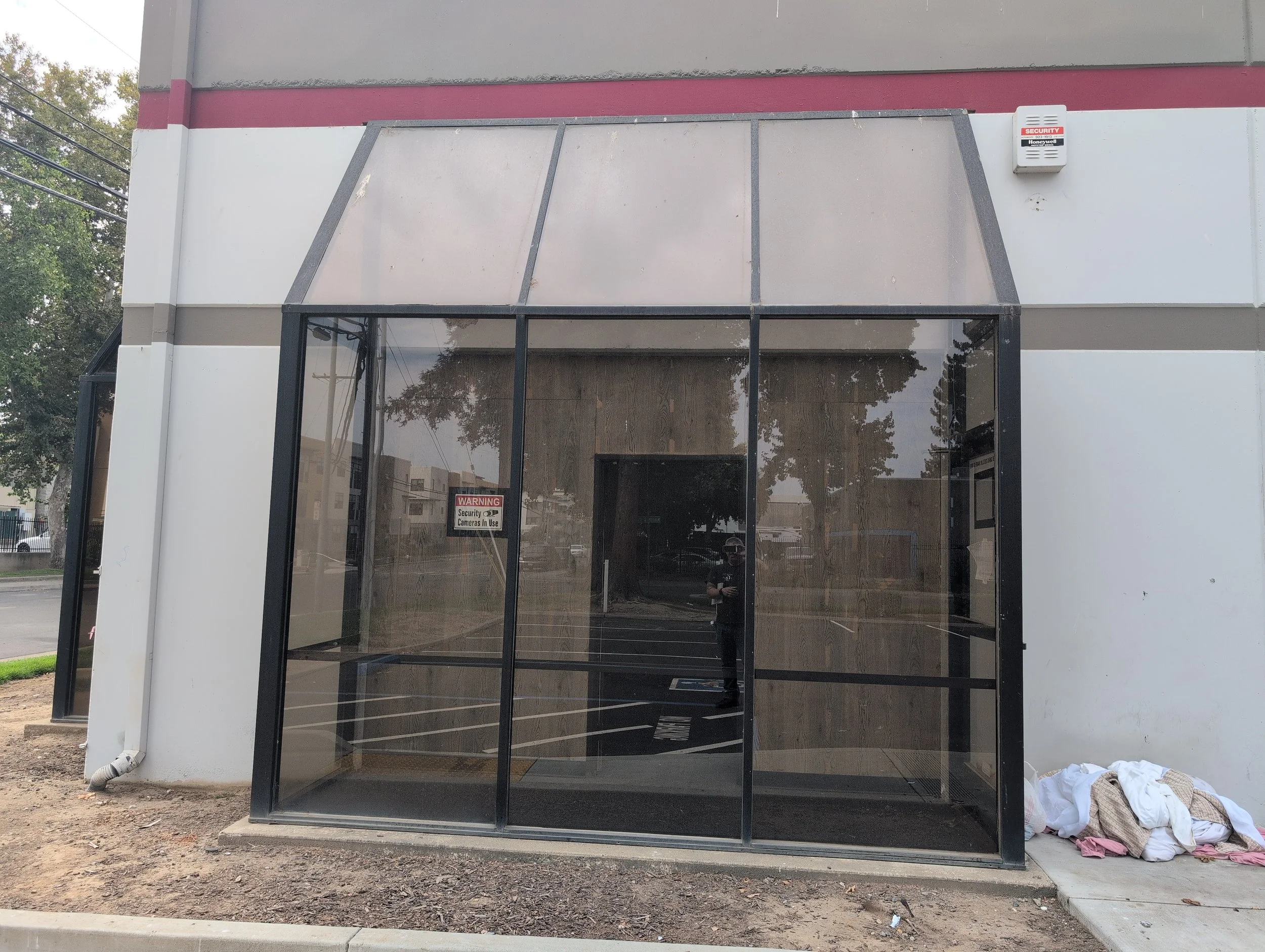 Empty storefront with glass doors and windows, warning sign about security cameras, and some laundry or fabric on the sidewalk outside.