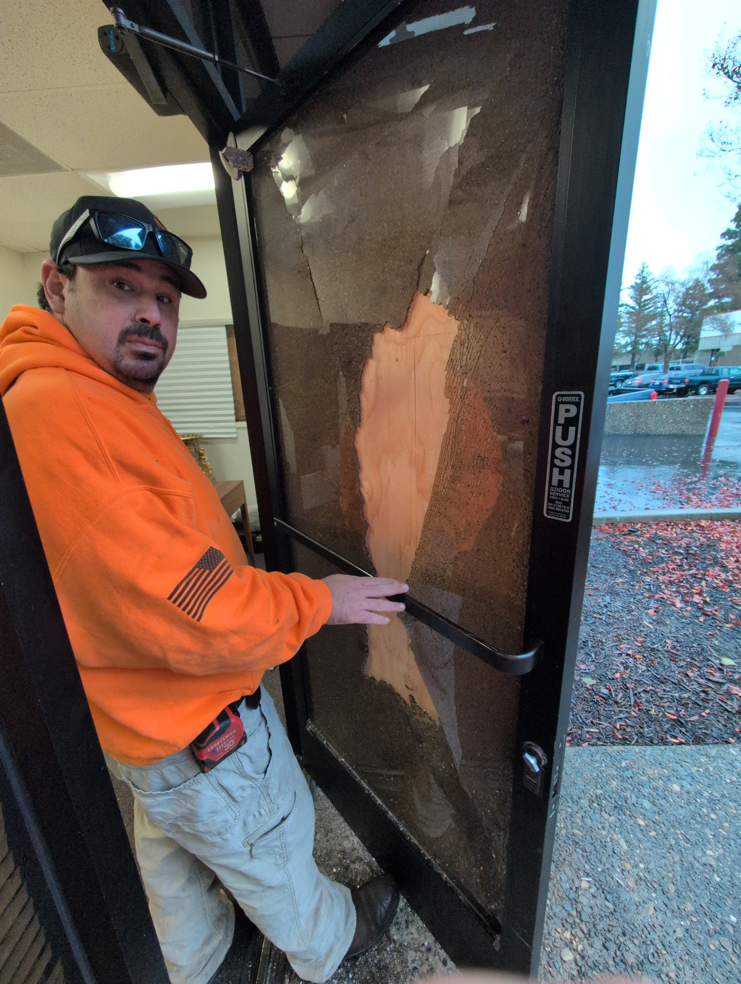 A man wearing an orange hoodie, sunglasses on his cap, and beige shorts is standing near a large glass display case with a cracked surface. The case contains a large wooden slab with natural wood grain patterns. Outside, there are parked cars and trees in the background.