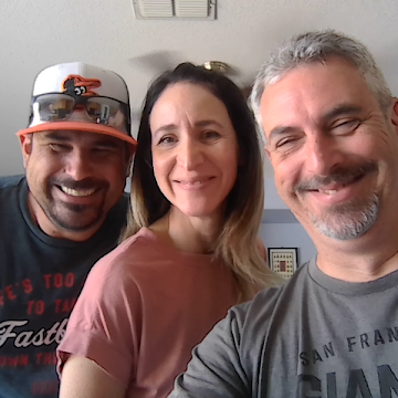 Three people smiling for a selfie indoors, with a man on the right in a gray shirt, woman in the center with long hair, and man on the left wearing a baseball cap.