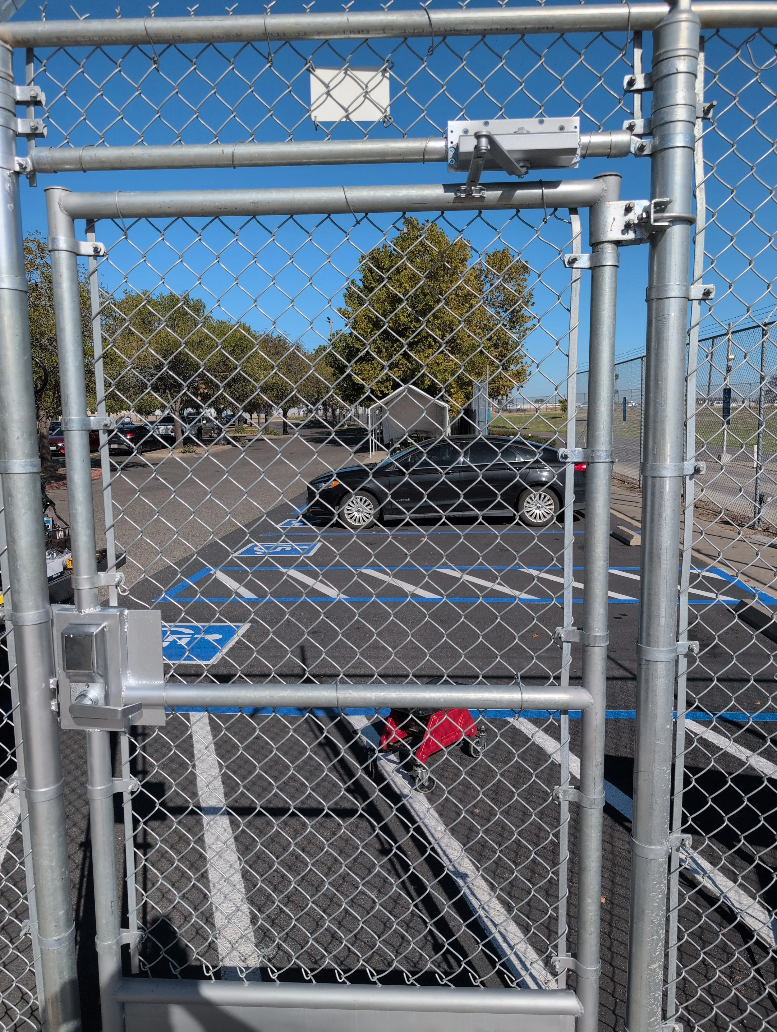 A parking lot seen through a metal chain-link gate with a lock, with trees and parked cars in the background, and a small structure visible beyond the lot.